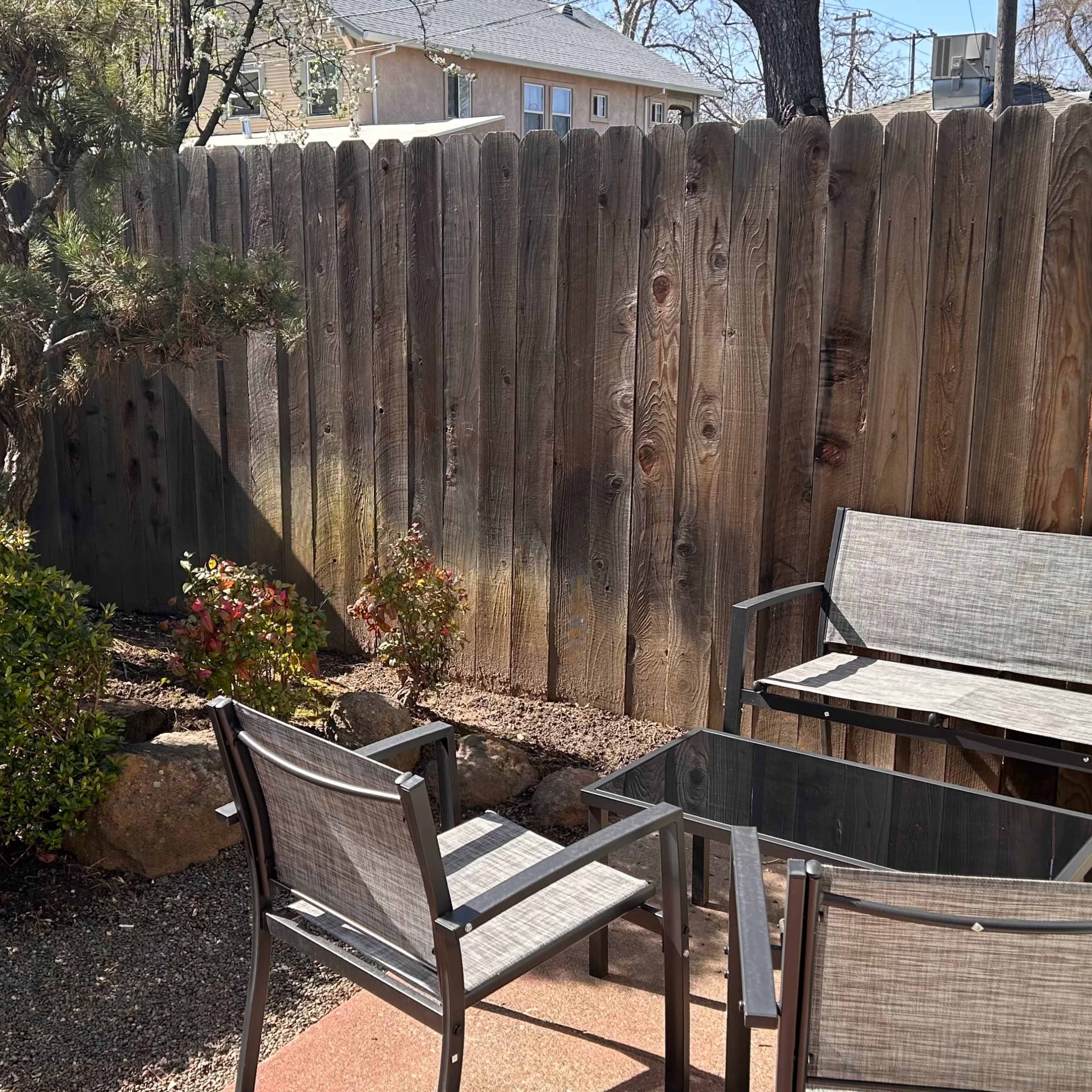 A small outdoor patio features two chairs and a table surrounded by a bush and a tree, enclosed by a wooden fence.