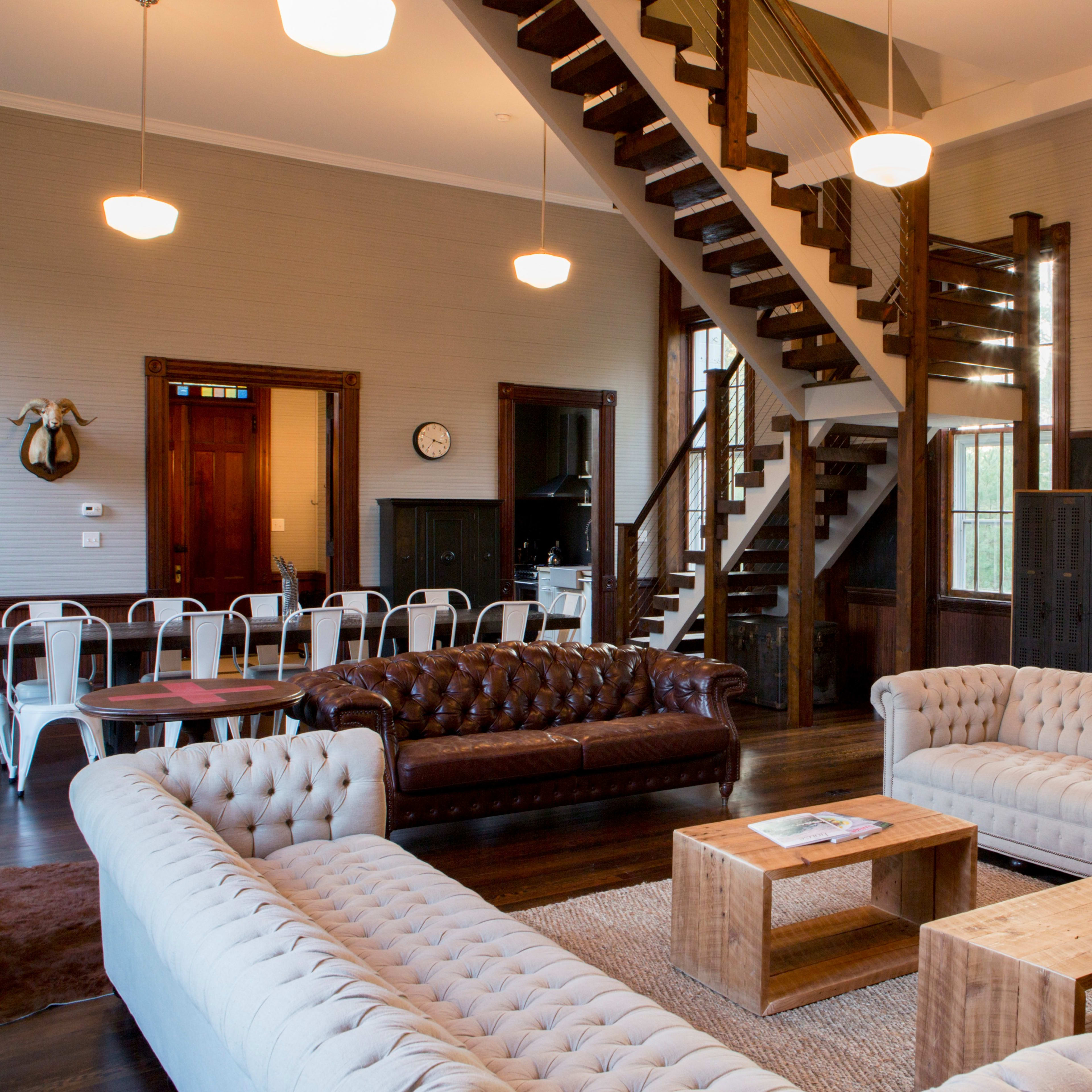 The room features a spacious interior with two tufted sofas, wooden furniture, and a staircase leading to an upper level.