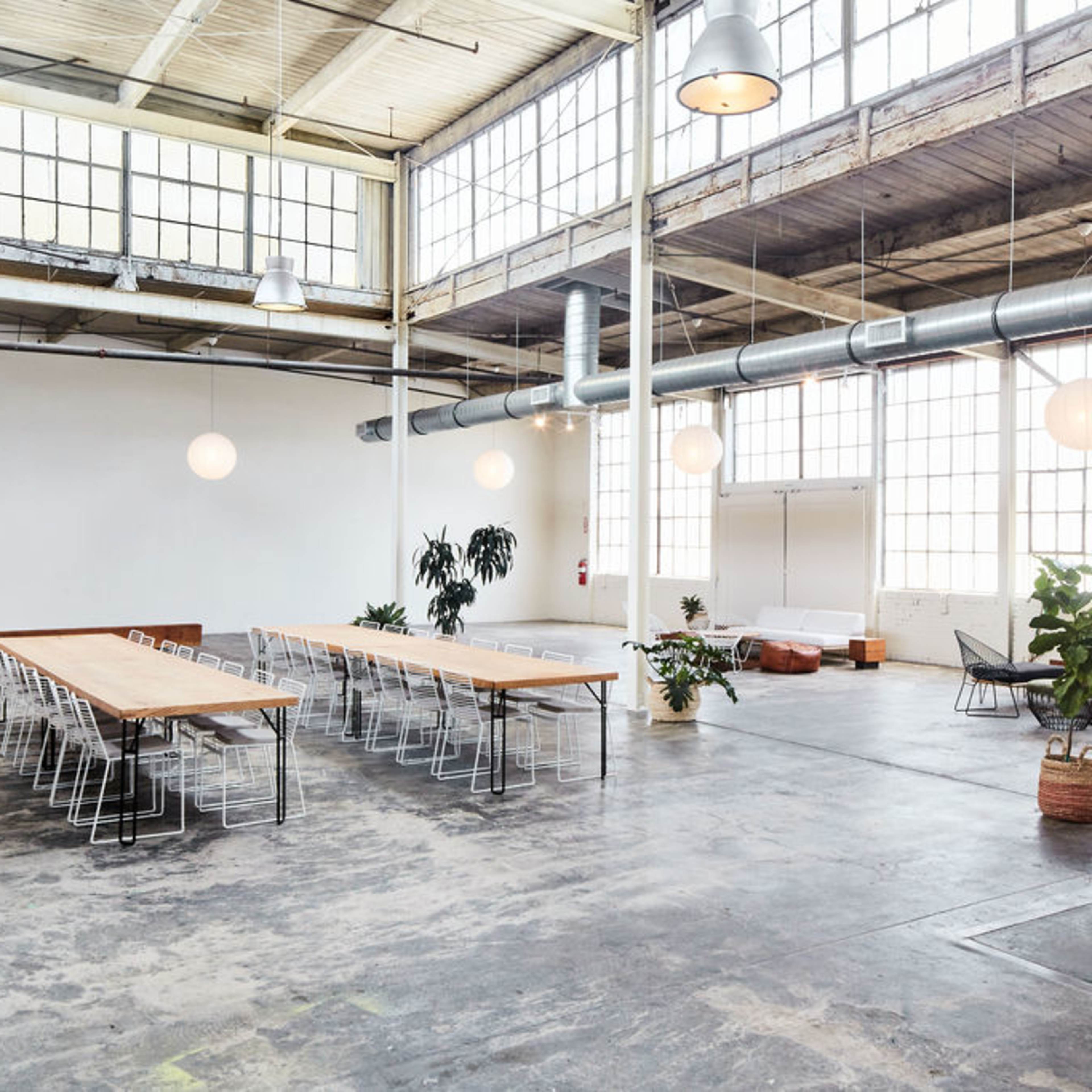 A spacious, industrial-style room with large windows, a long wooden table surrounded by metal chairs, and various plants positioned around the area.