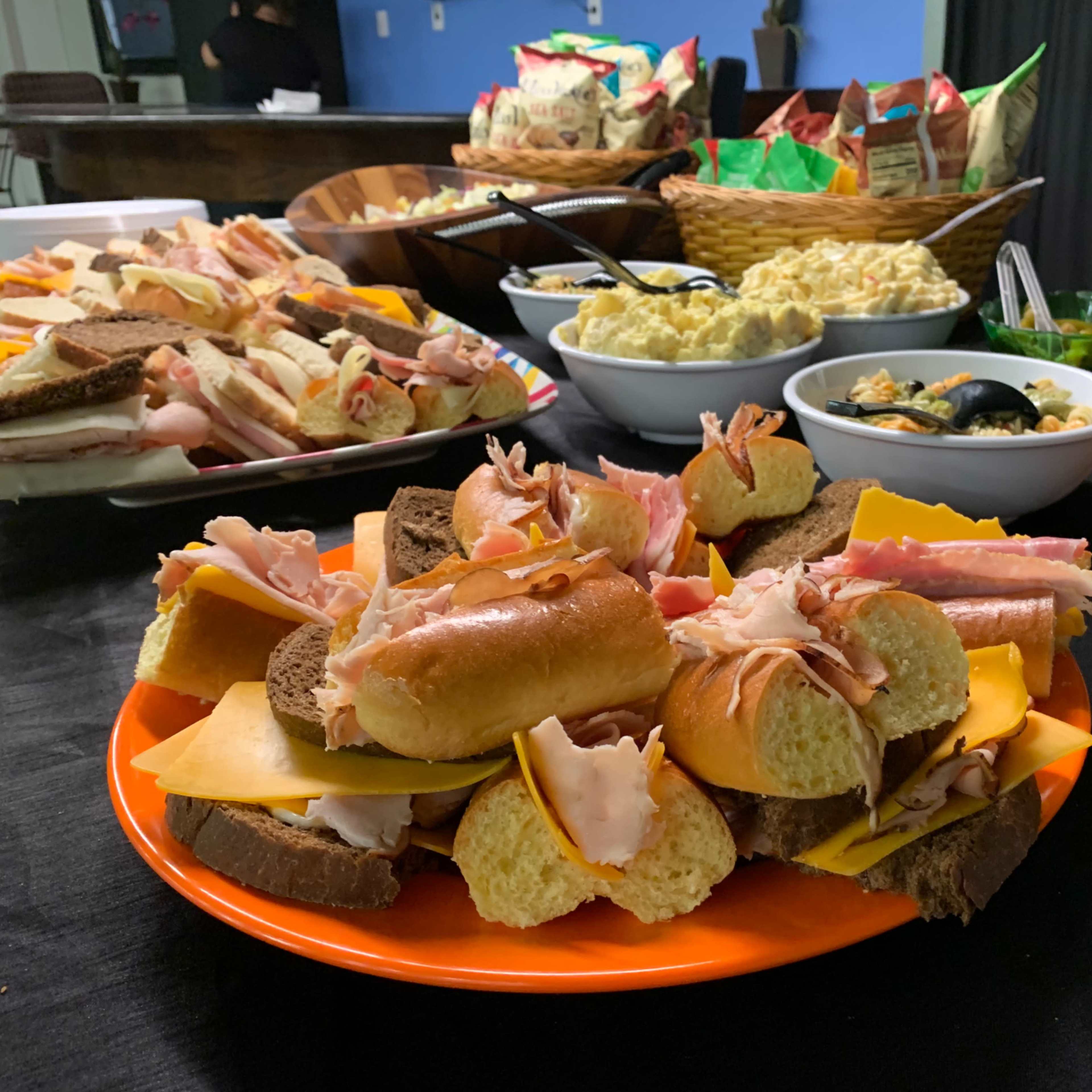 A platter of assorted sandwiches and meats is displayed on an orange plate, surrounded by bowls of pasta salad, vegetable dishes, and snacks on a table.