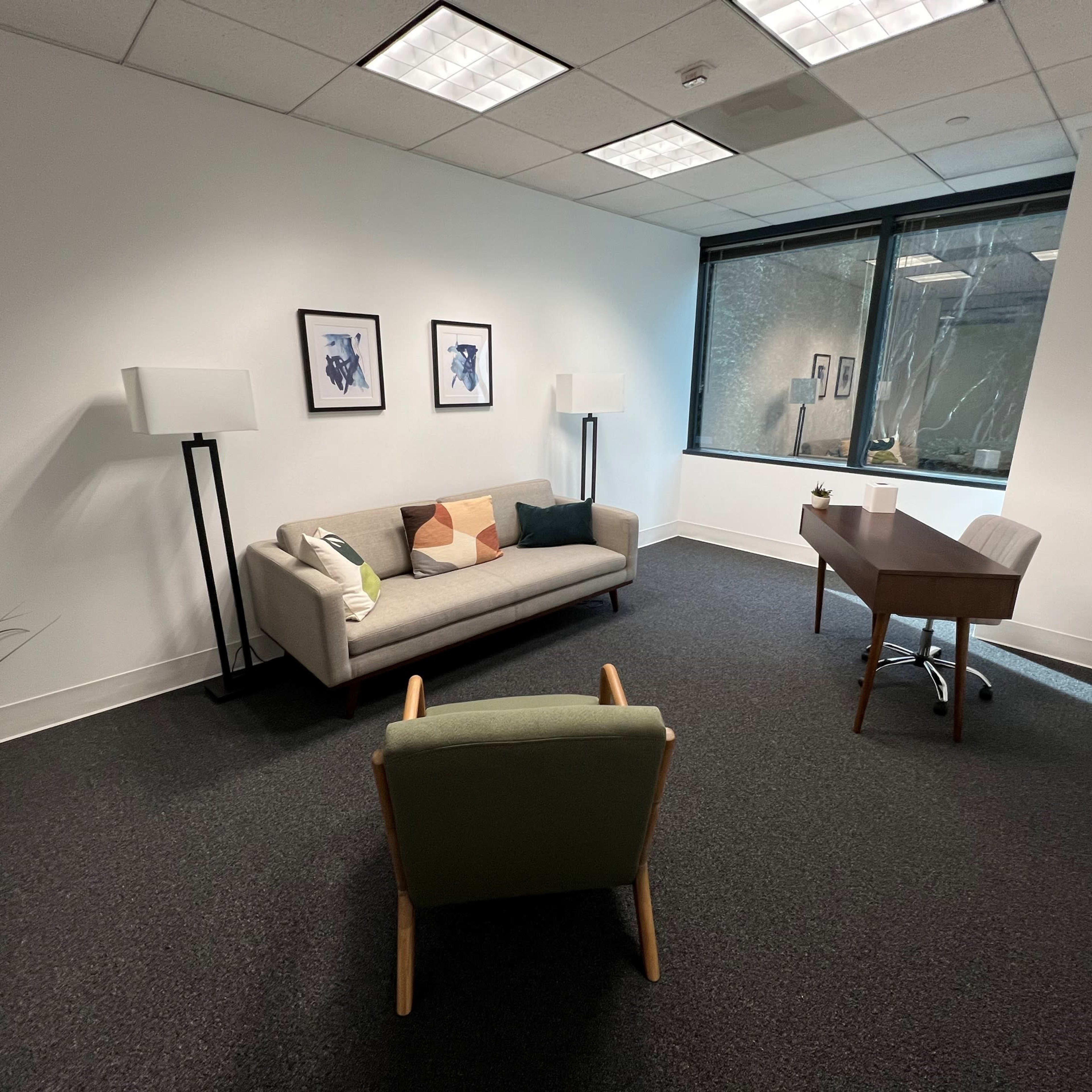 A minimalist office space featuring a gray sofa, a wooden desk with a chair, and two floor lamps against a white wall with two framed pictures.