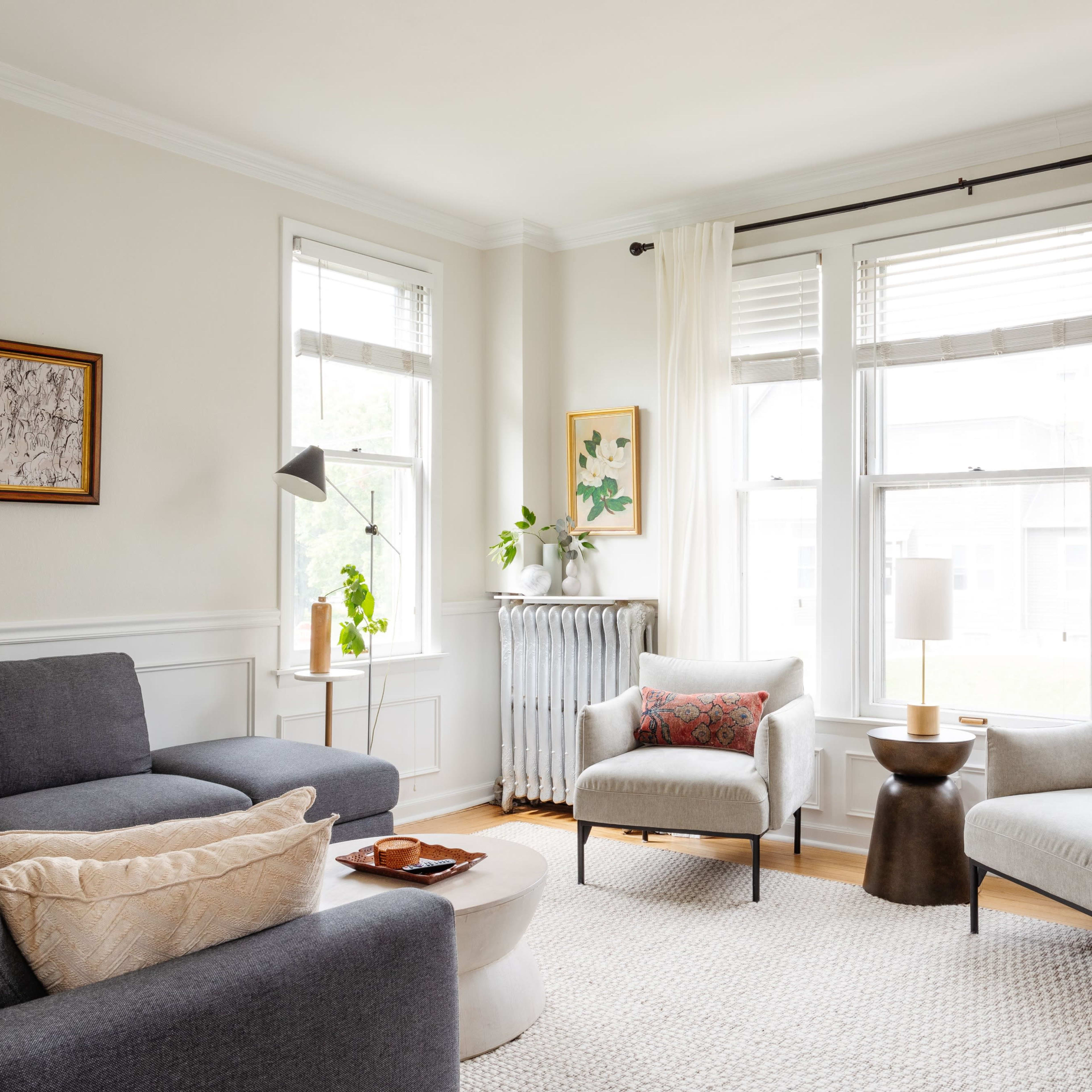 A bright living room with a gray sofa, two light-colored armchairs, a round coffee table, and large windows letting in natural light.