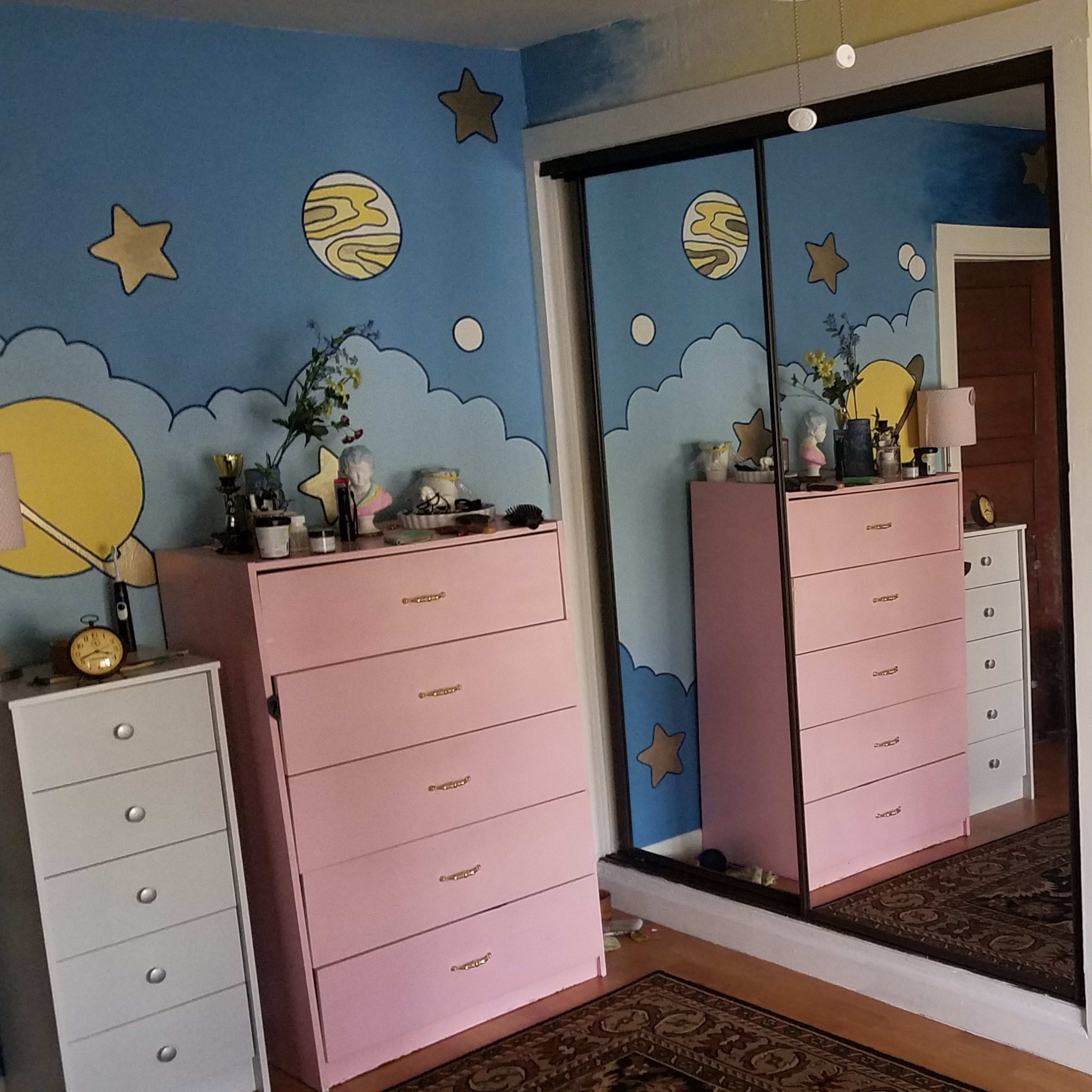 A brightly colored bedroom featuring blue walls with cloud and celestial-themed murals, a pink dresser, and a mirrored closet.