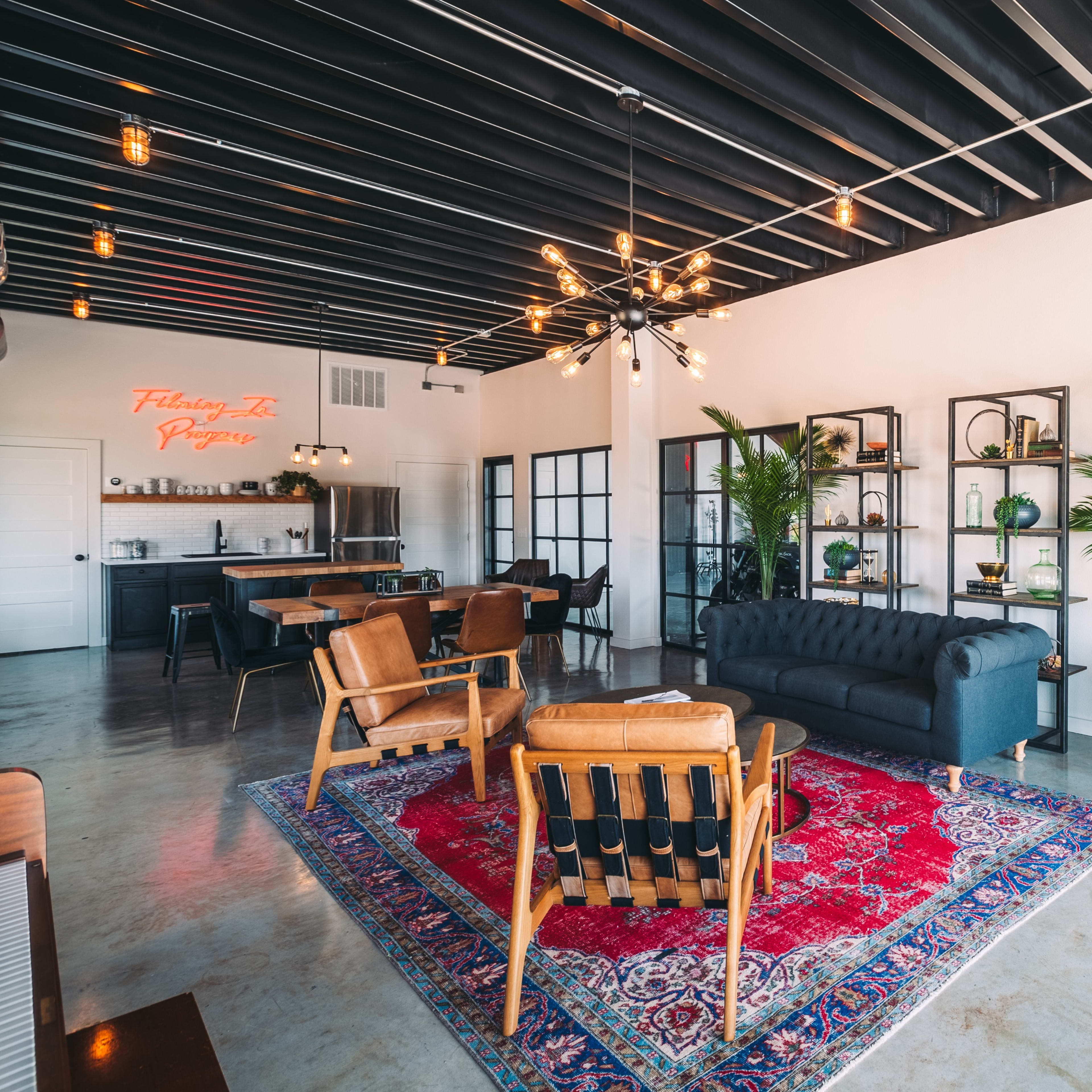 A modern, open interior space with a mix of seating areas, decorative plants, and shelves, featuring a large rug and unique light fixtures.
