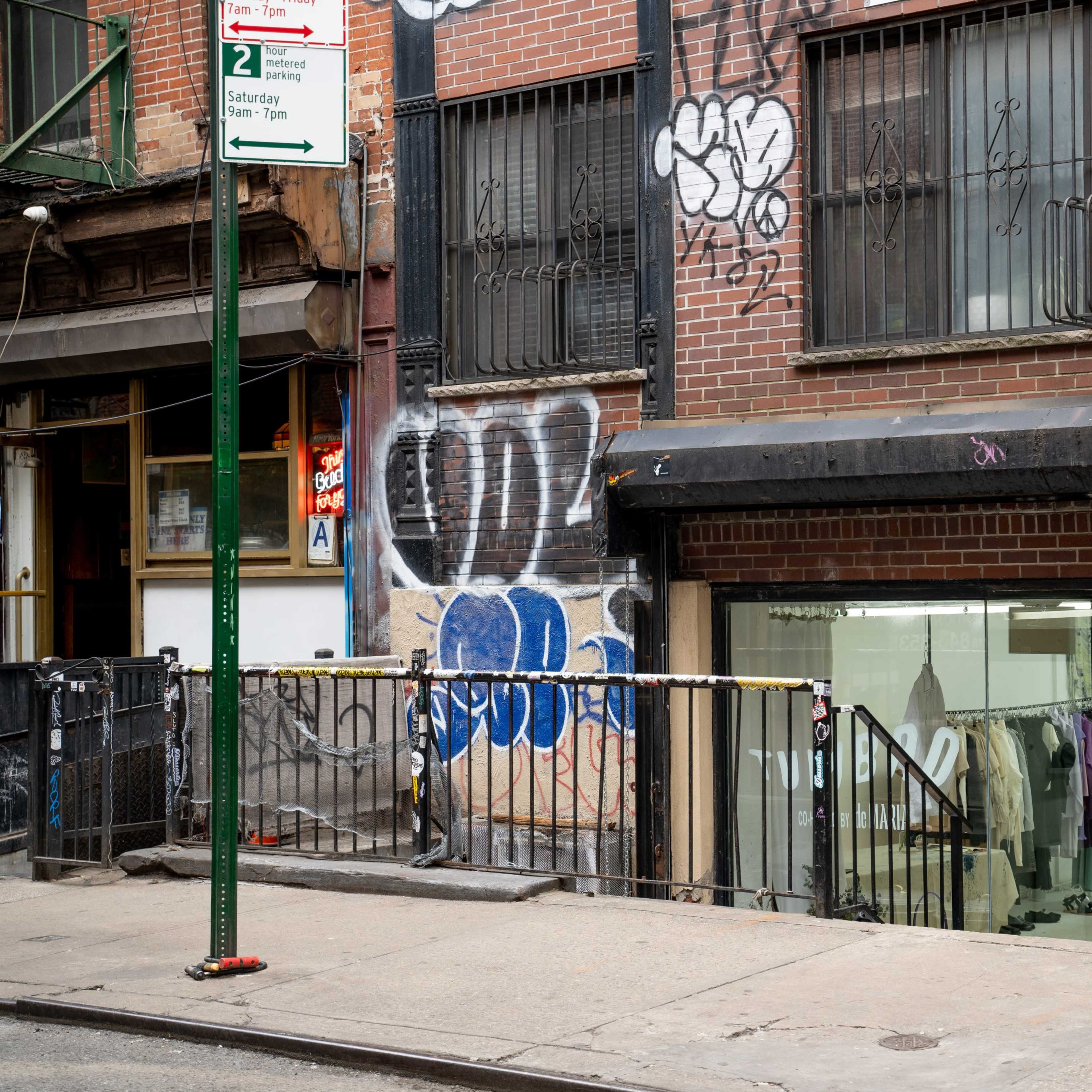 A brick building with graffiti on the exterior features a staircase leading up to a glass storefront entrance.