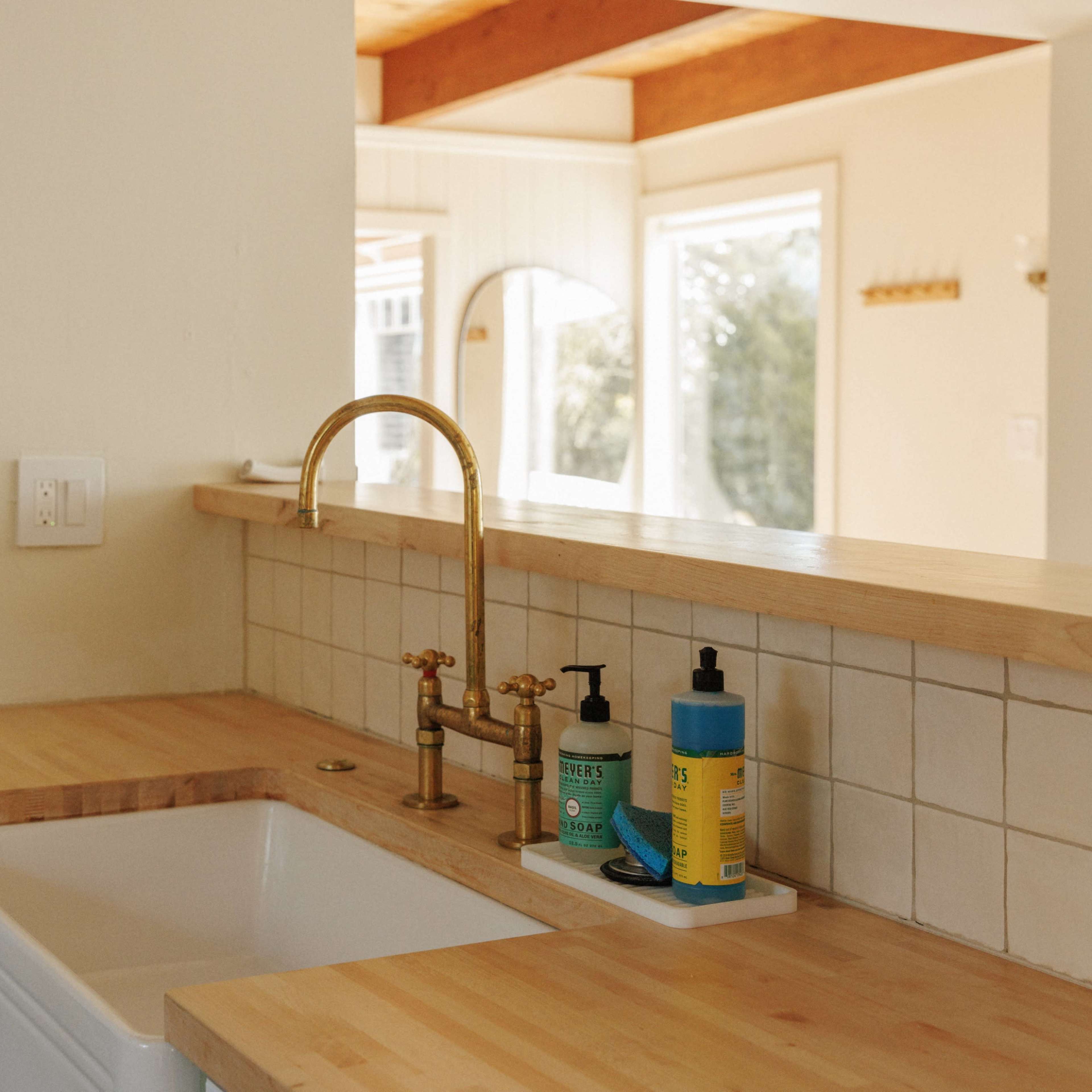 A kitchen counter with a farmhouse sink, a gold faucet, and various cleaning products arranged on a light-colored wood surface.