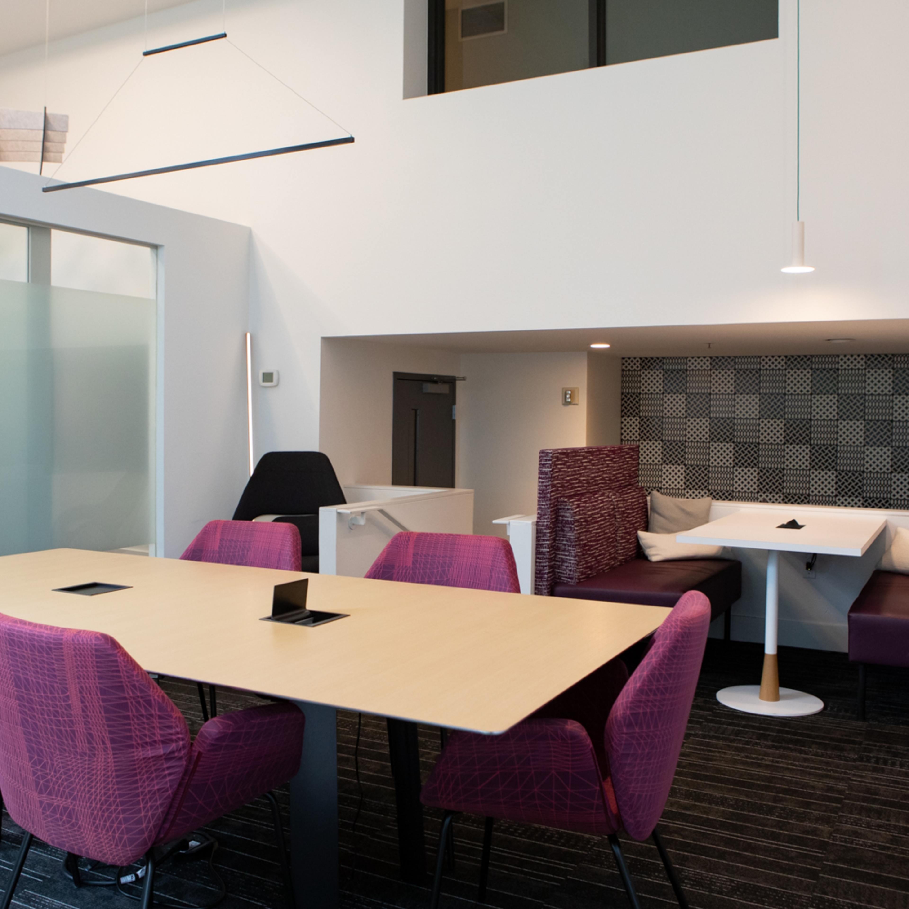 A modern office space featuring a conference-style table surrounded by pink chairs, with a booth area and a wall that includes a graphic design.