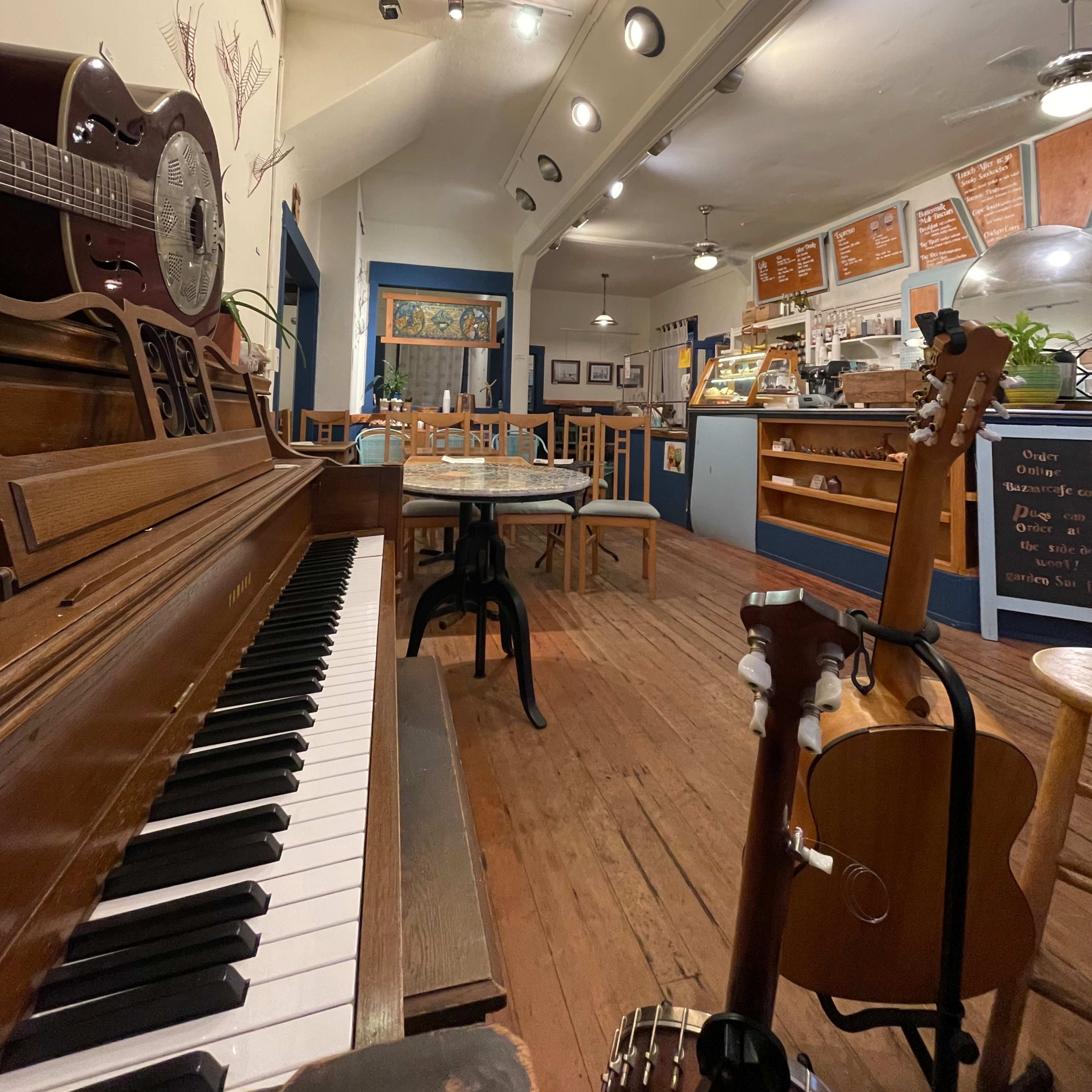 A cozy cafe interior featuring a wooden piano, tables, and guitars, with a counter displaying a menu on the back wall.