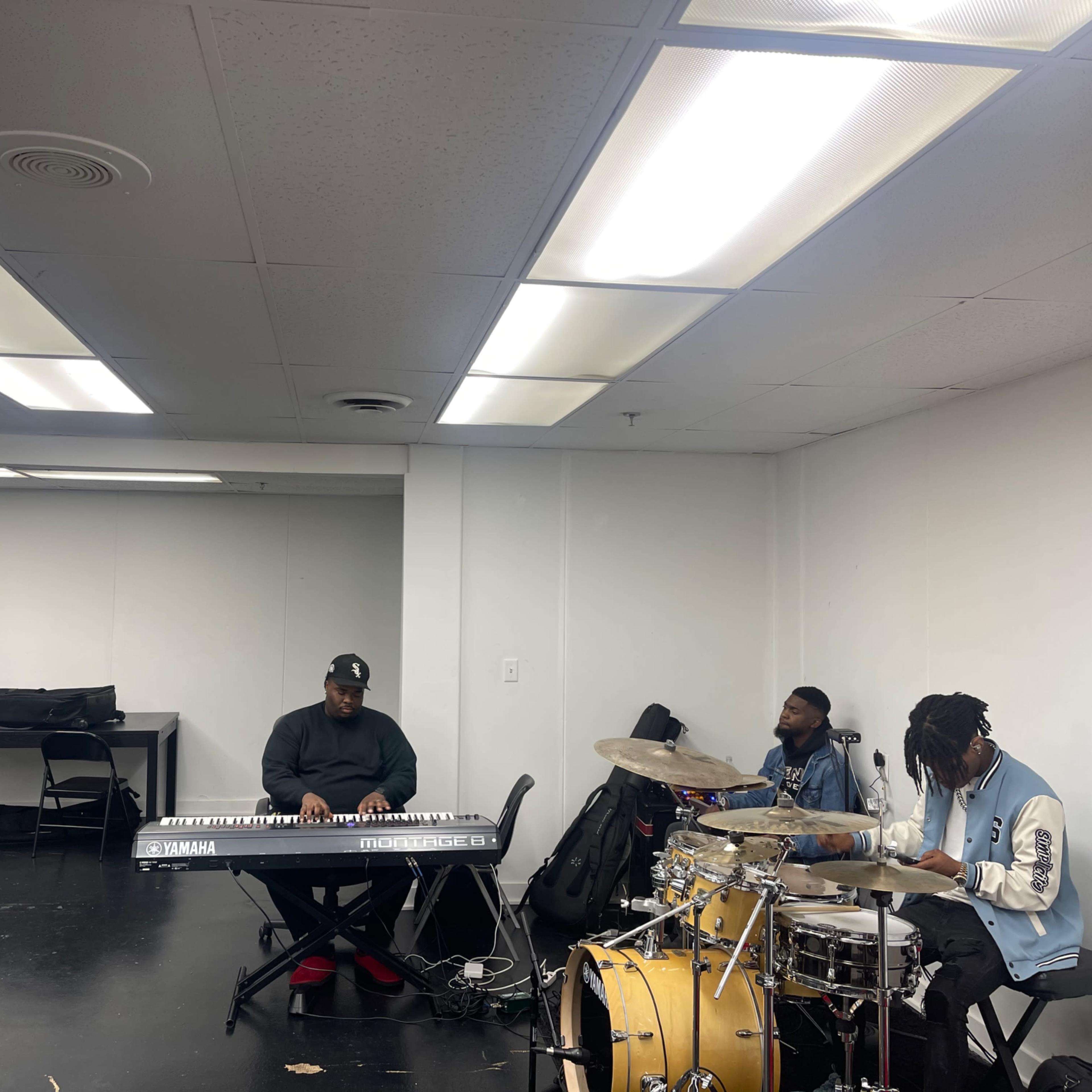 Three musicians are rehearsing in a spacious, well-lit room, with one playing a keyboard, and two others on drums and percussion.