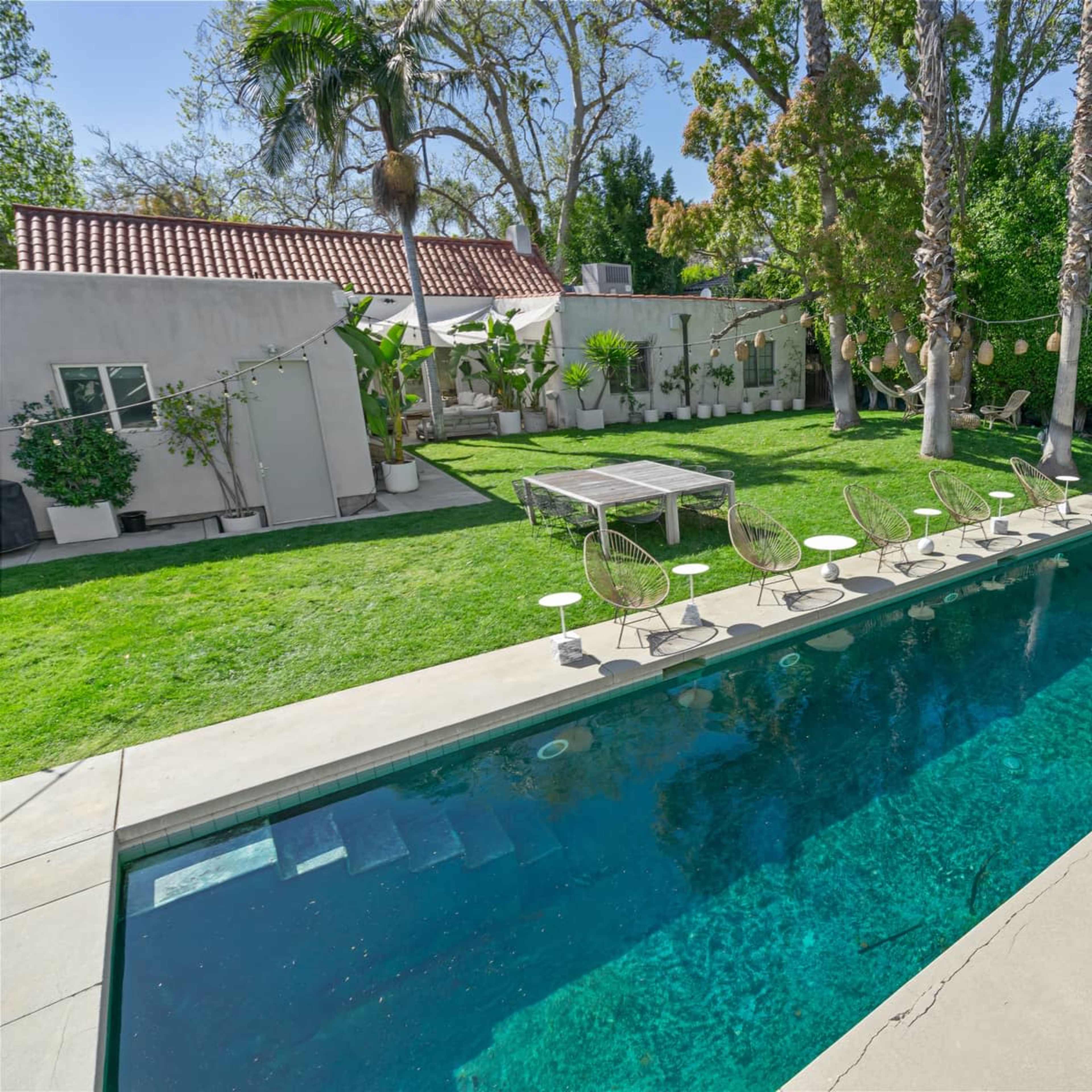 A backyard with a swimming pool surrounded by green grass, a patio table, and several chairs, flanked by trees and decorative plants.