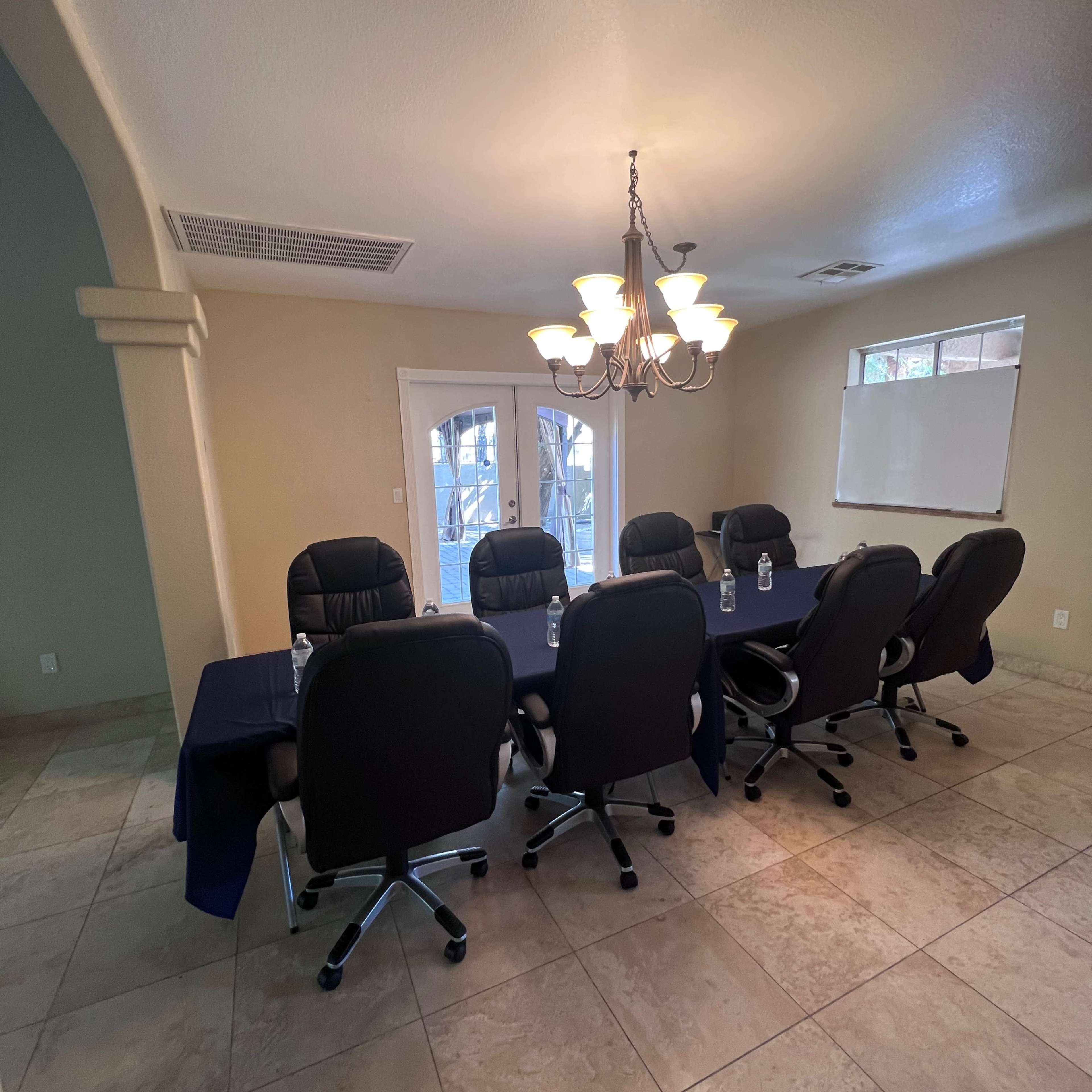 A conference room features a long table surrounded by six black office chairs, with a chandelier overhead and large windows providing natural light.