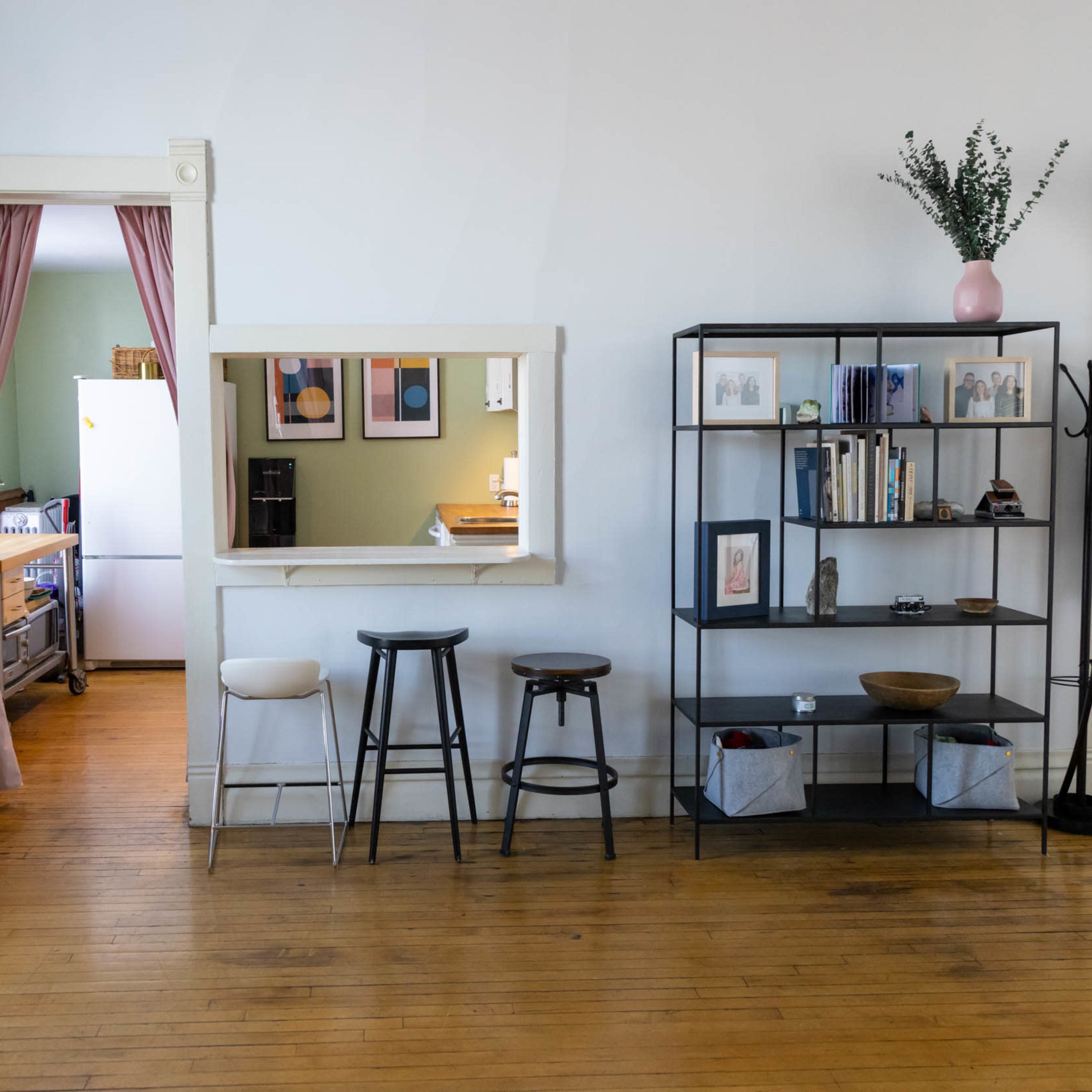 A spacious room with a view of a doorway leading to a kitchen area, a shelf with books and decor, and several stools lined up against a wall.
