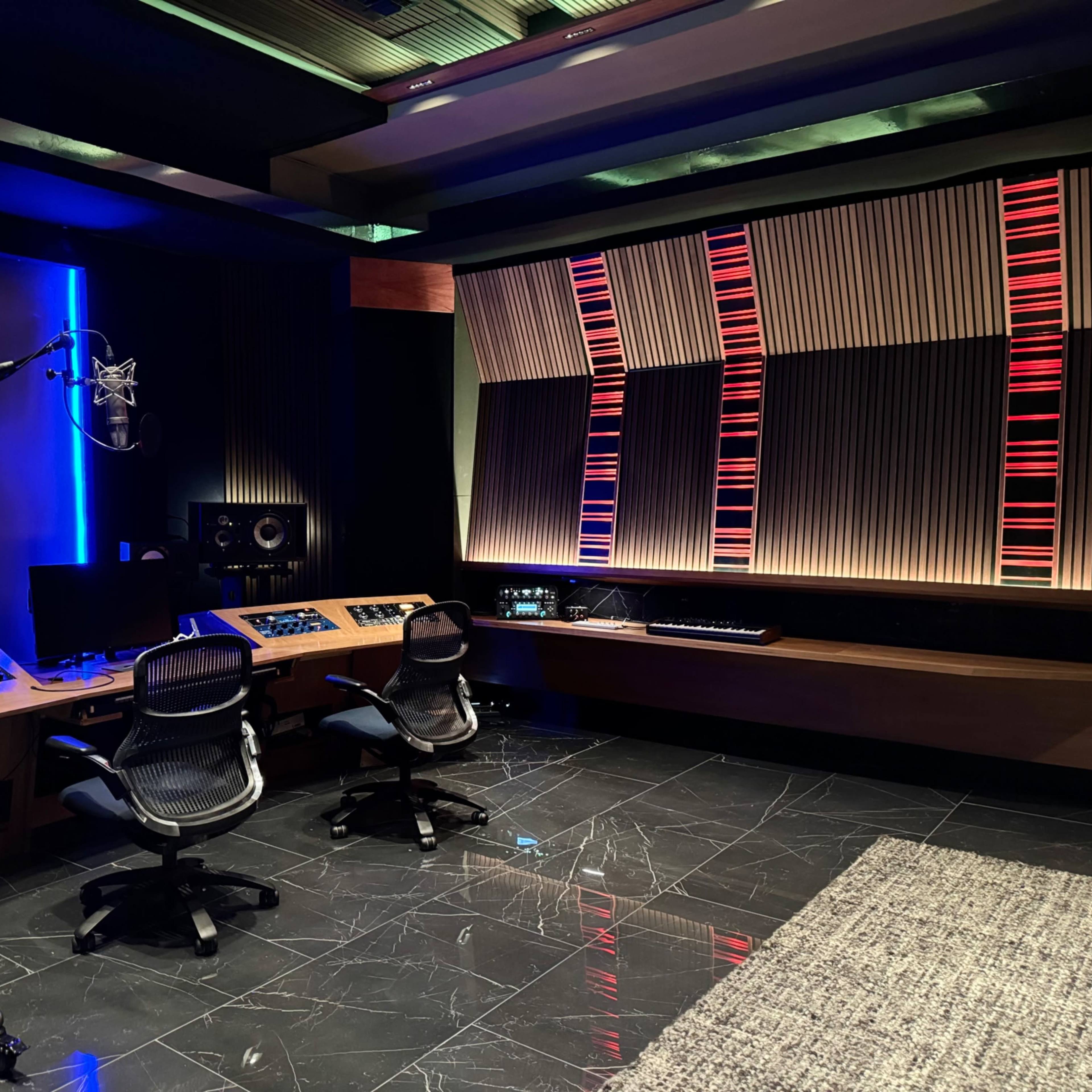 A modern music recording studio featuring a microphone, sound mixing equipment, and two chairs facing a soundboard, with a large screen and illuminated panels on the walls.