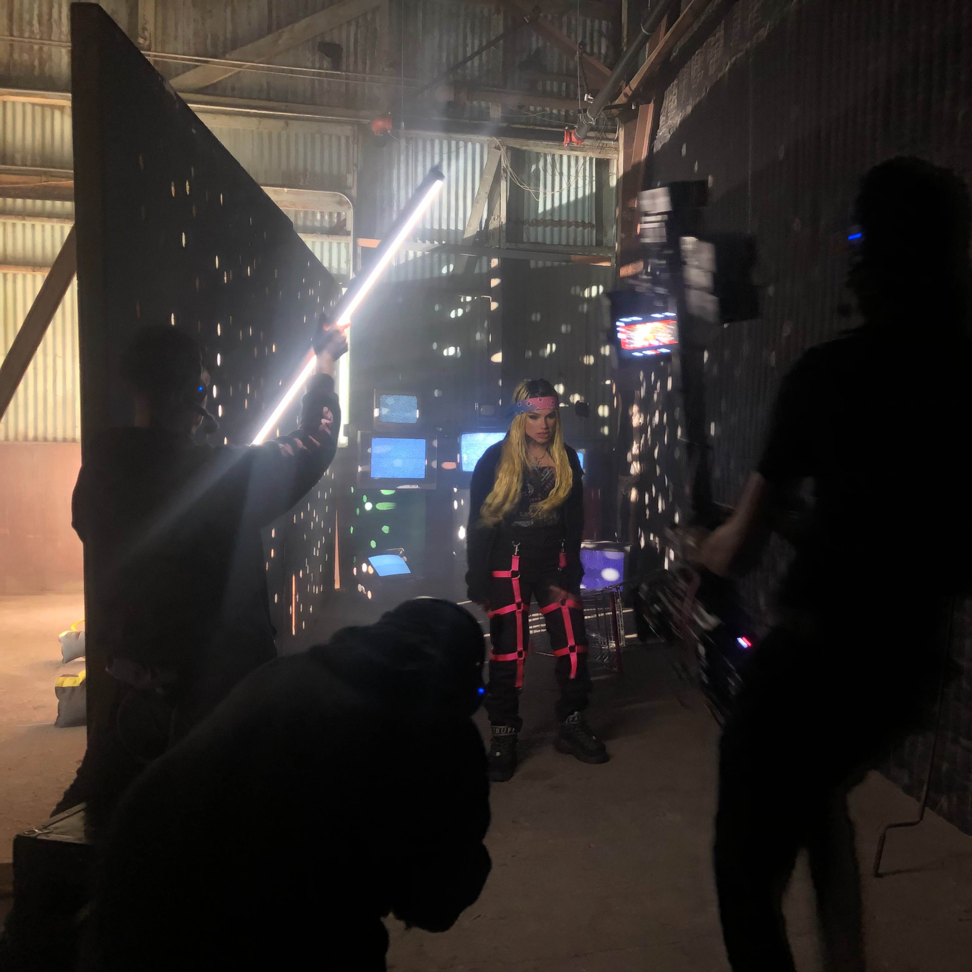 A person with long hair stands in a dimly lit industrial space surrounded by illuminated screens and people positioning lights and cameras.