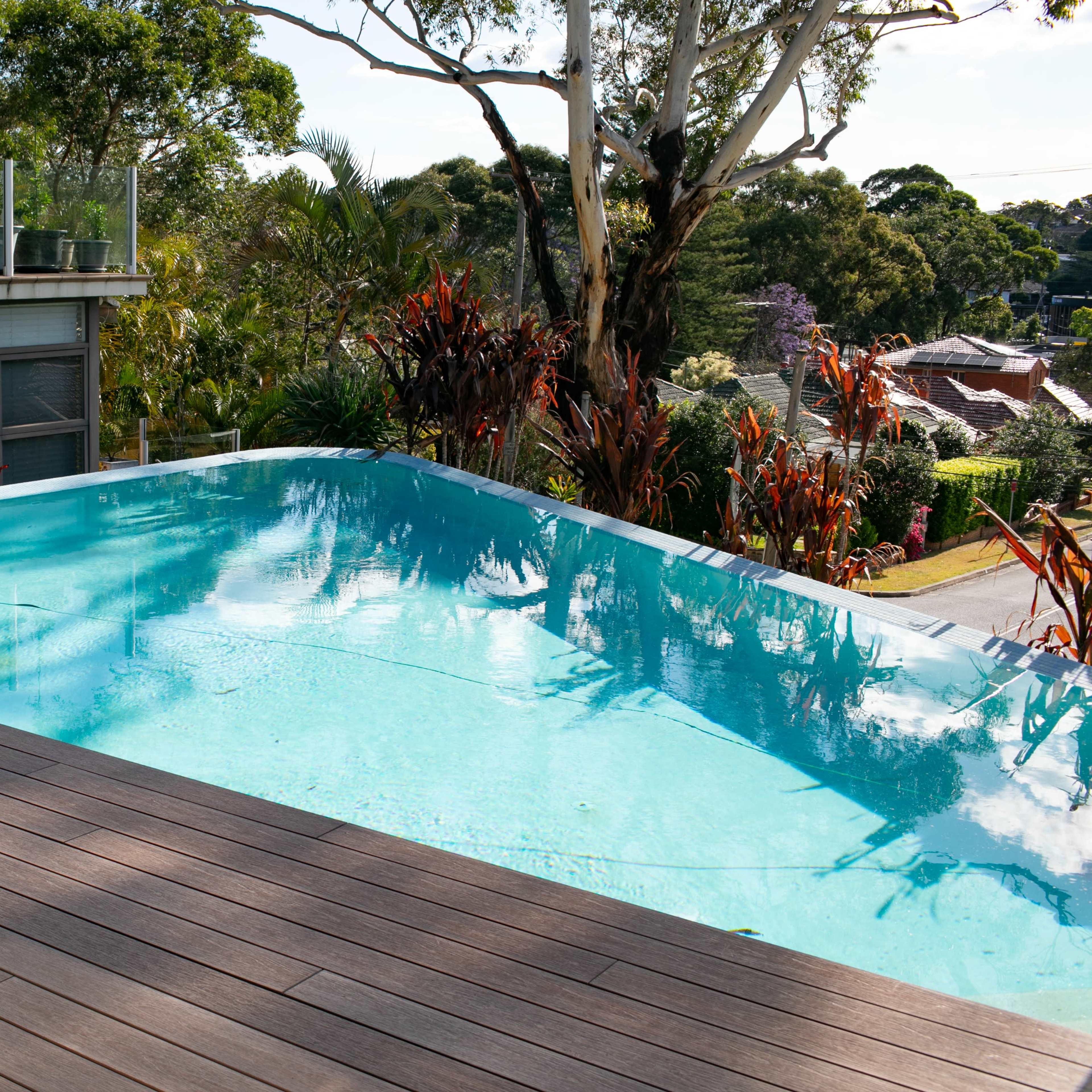 A modern infinity pool adjacent to a wooden deck, surrounded by lush greenery and residential houses in the background.