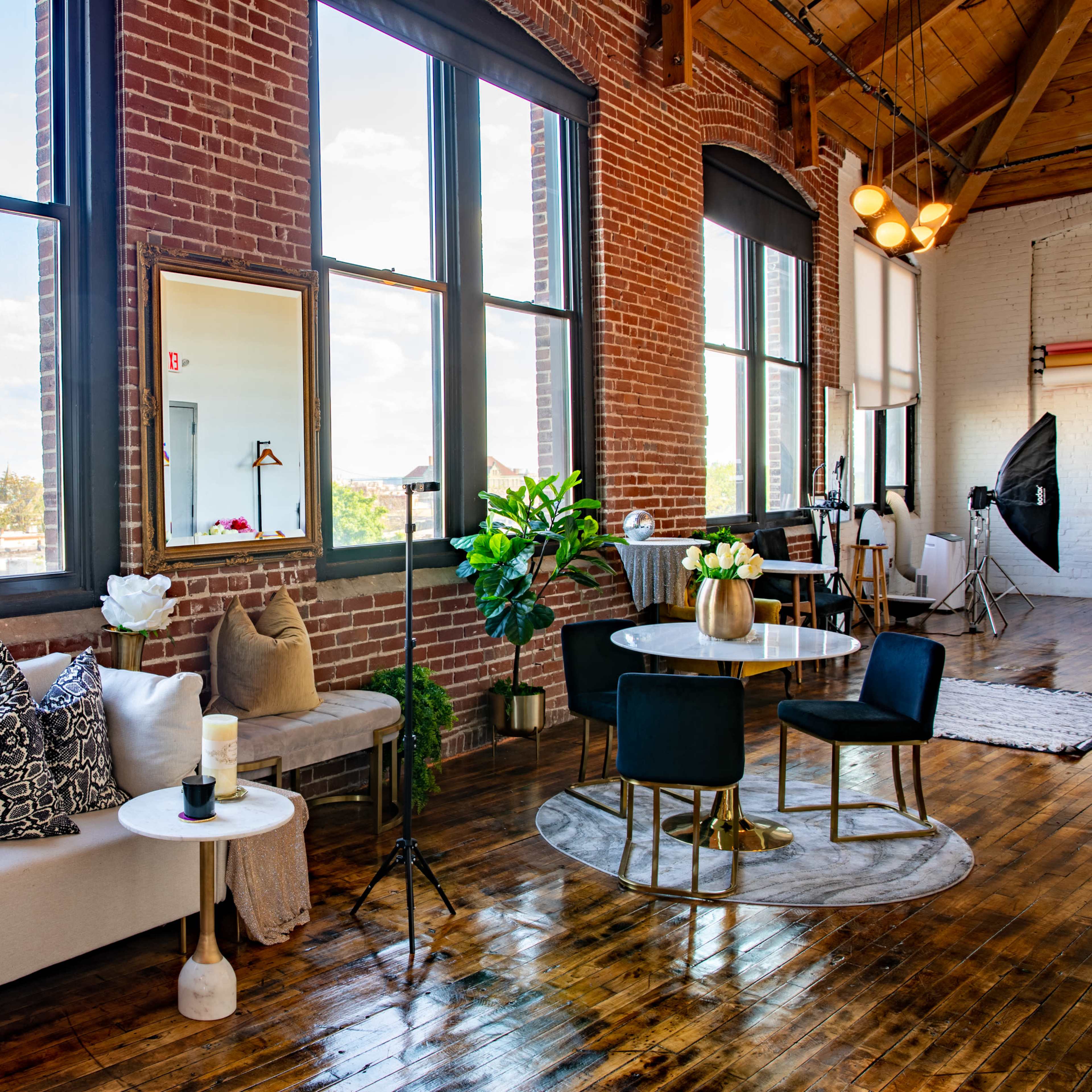 A bright urban loft features large windows, exposed brick walls, a cozy sofa with decorative pillows, a round dining table, and a potted plant.