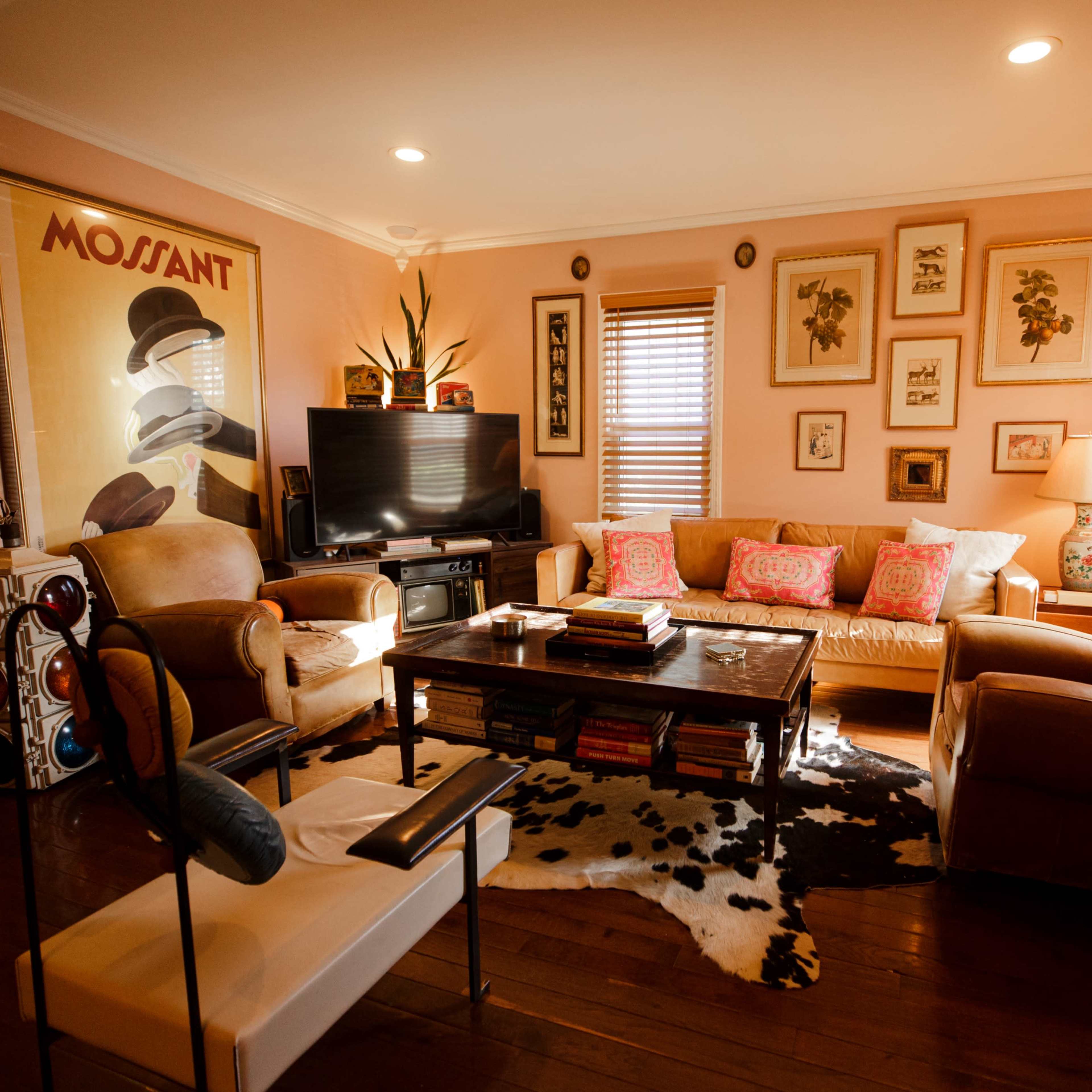The living room features a television, a coffee table stacked with books, and seating arranged around a cowhide rug, with framed artwork on the walls.