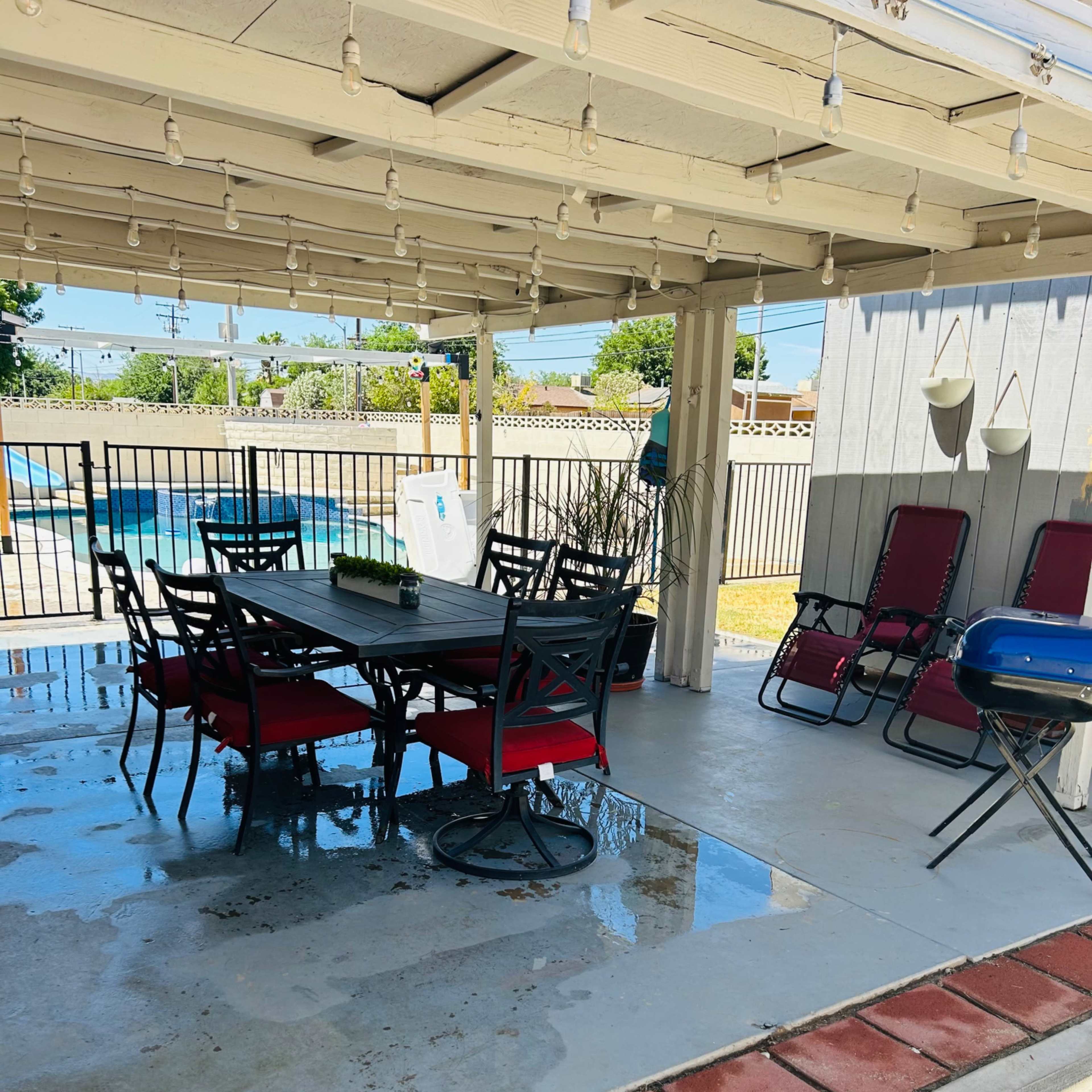 A covered patio features a table with chairs and a grill, overlooking a pool enclosed by a gate.