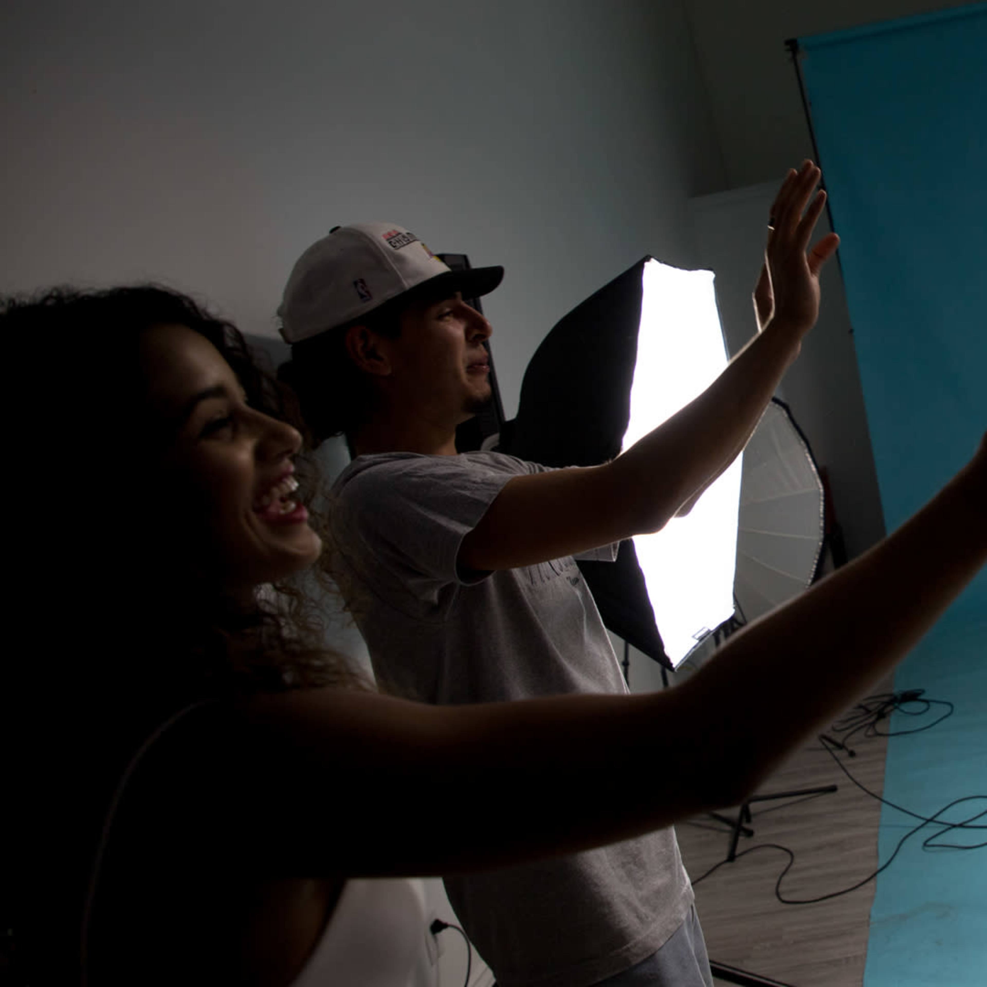 A performer raps into a microphone while two audience members engage enthusiastically, and a DJ manages the music at a photo shoot set with a blue backdrop.