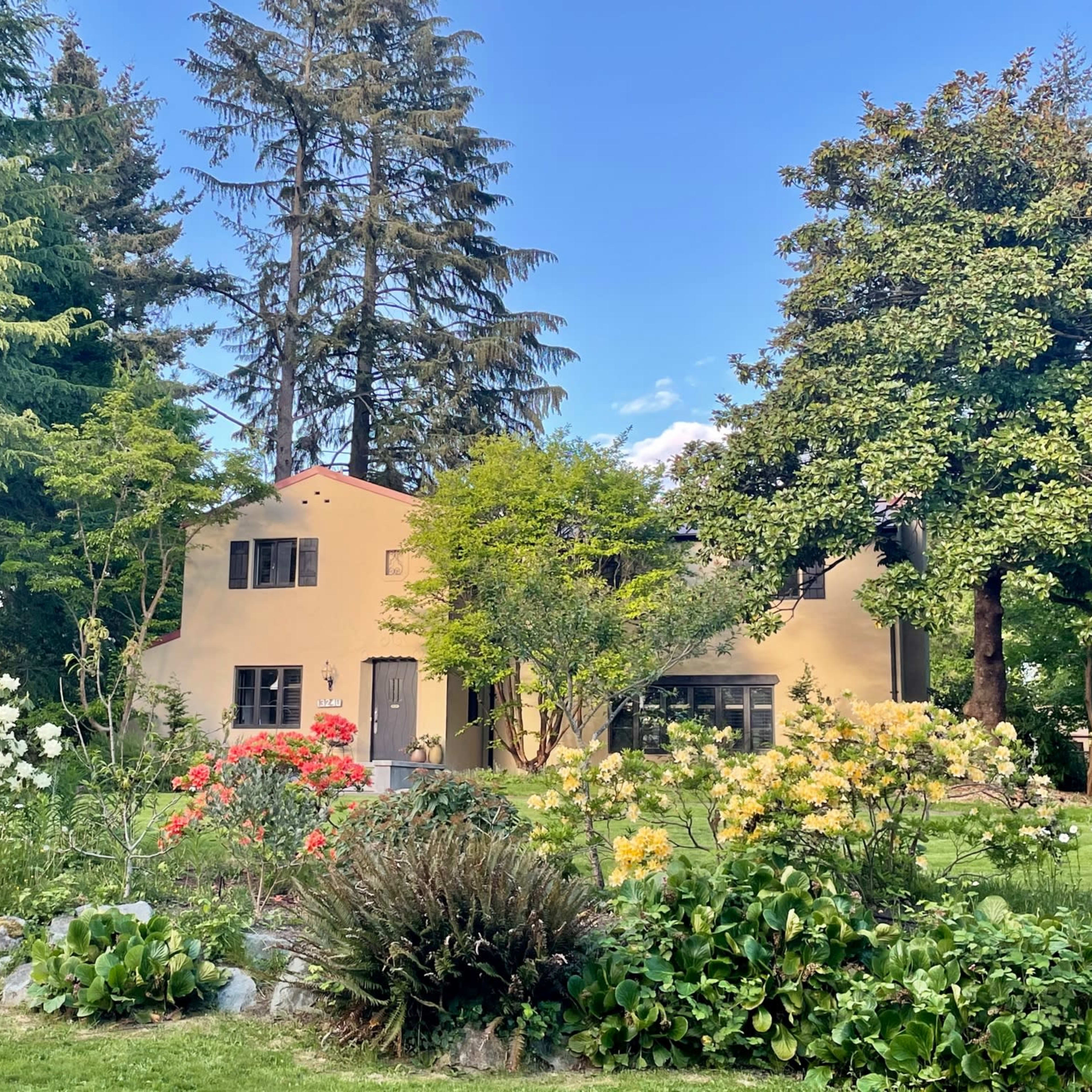 A two-story house with a light-colored exterior is surrounded by colorful flower gardens and tall trees.