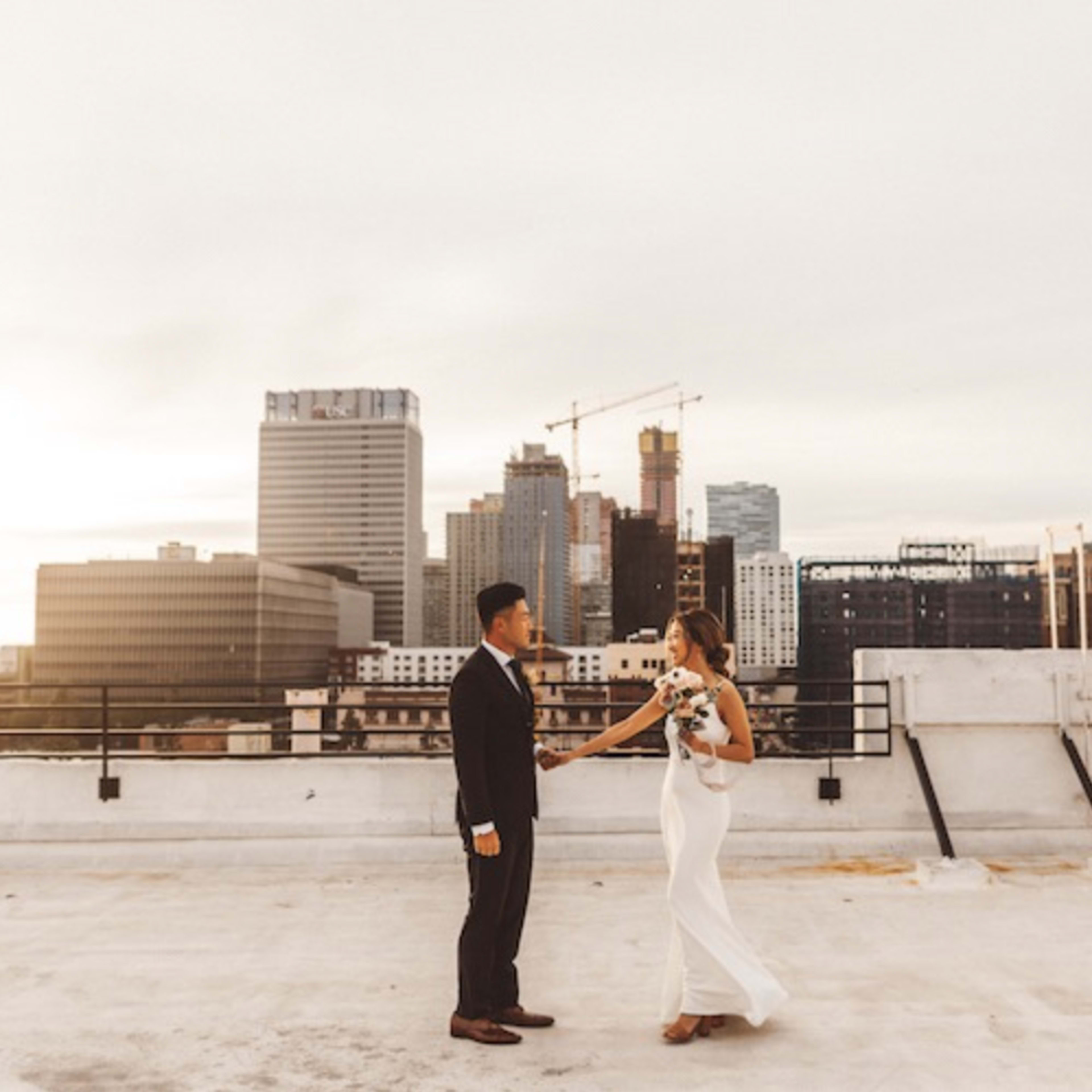 A couple stands on a rooftop at sunset, holding hands with a city skyline in the background.