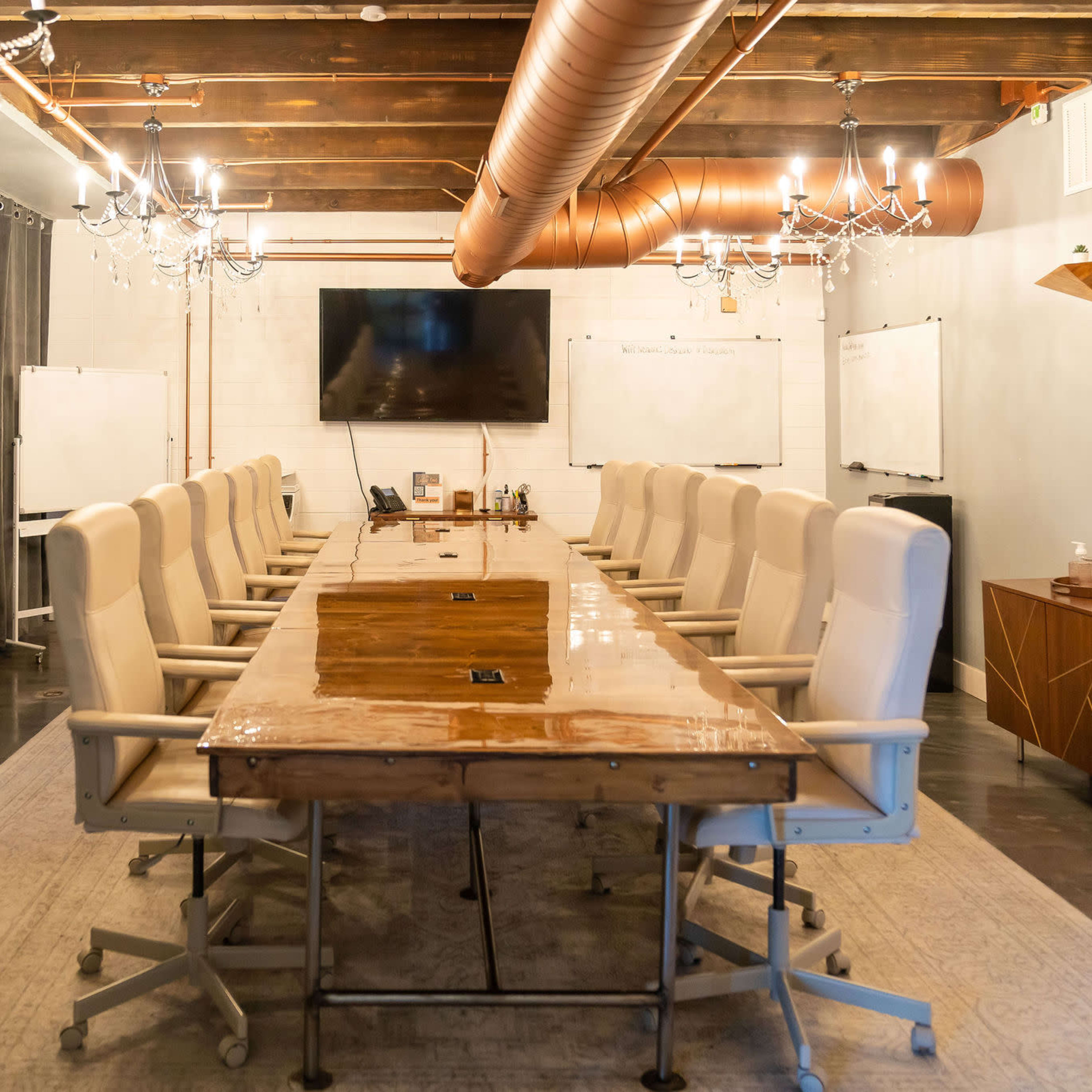 A conference room features a long wooden table surrounded by chairs, with a large screen on the wall and whiteboards on either side.