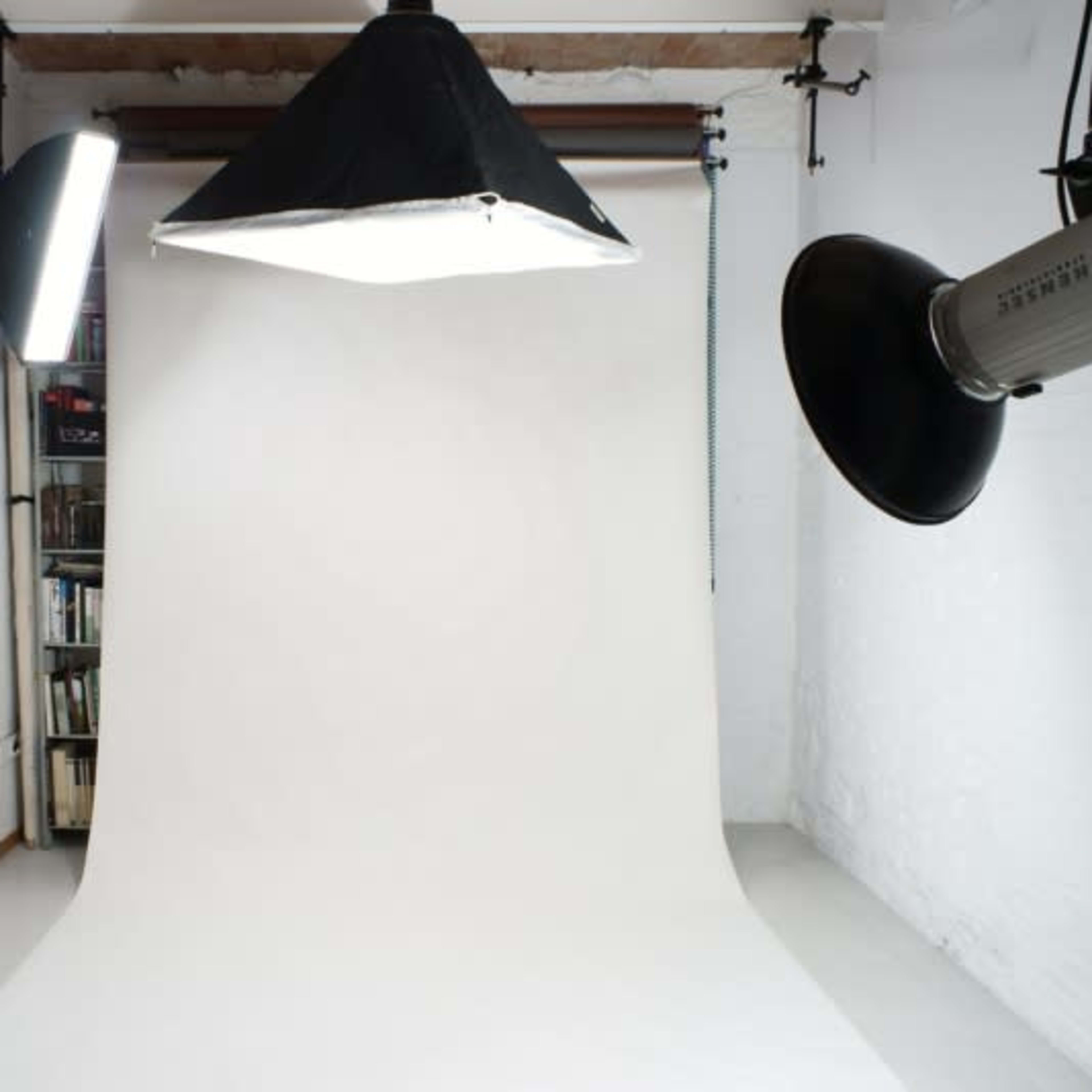 A photography studio with a white backdrop, overhead lighting, and a bookshelf in the background.
