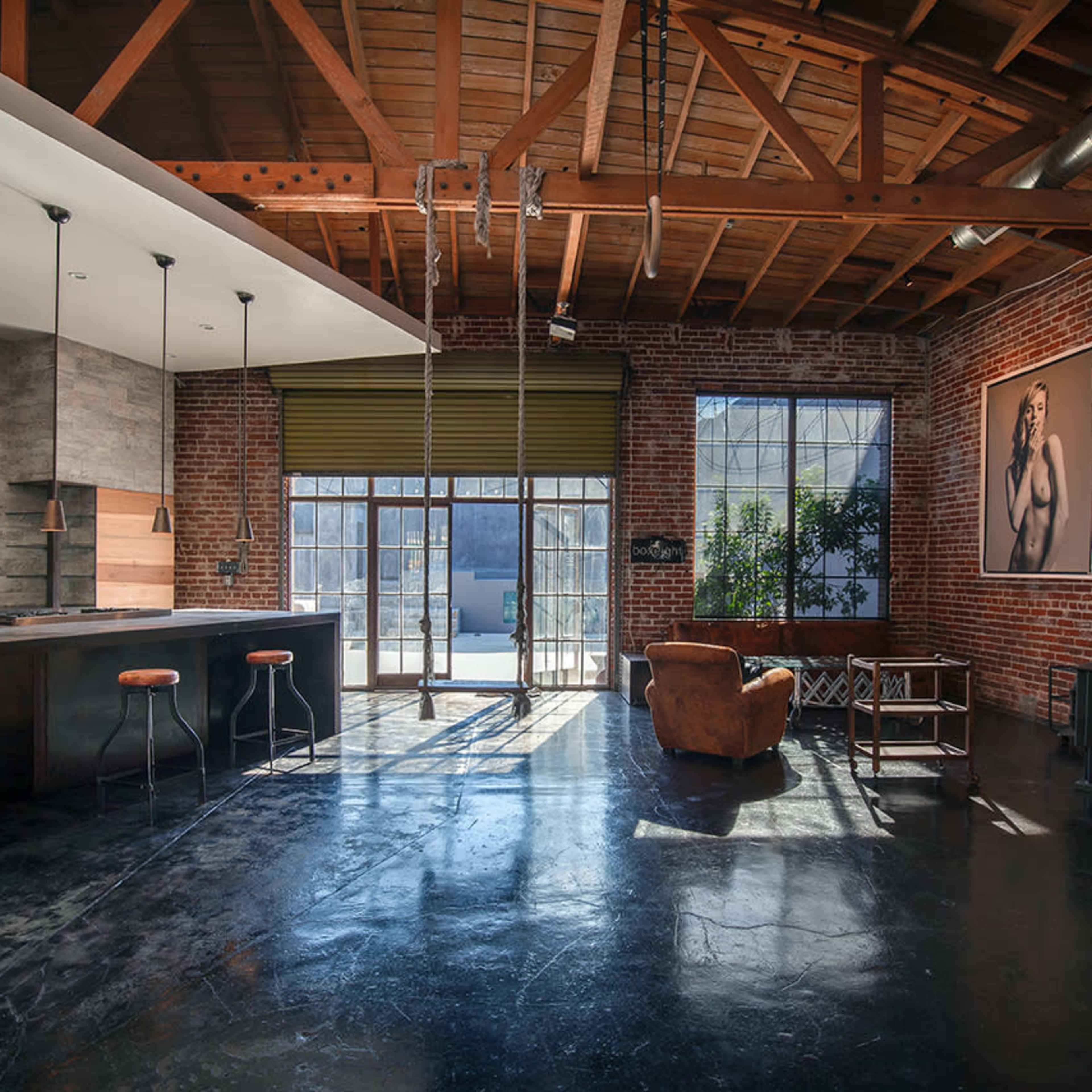 A spacious loft with exposed brick walls, industrial-style beams, a kitchen area with a dark countertop, and large windows that open to an outdoor space.
