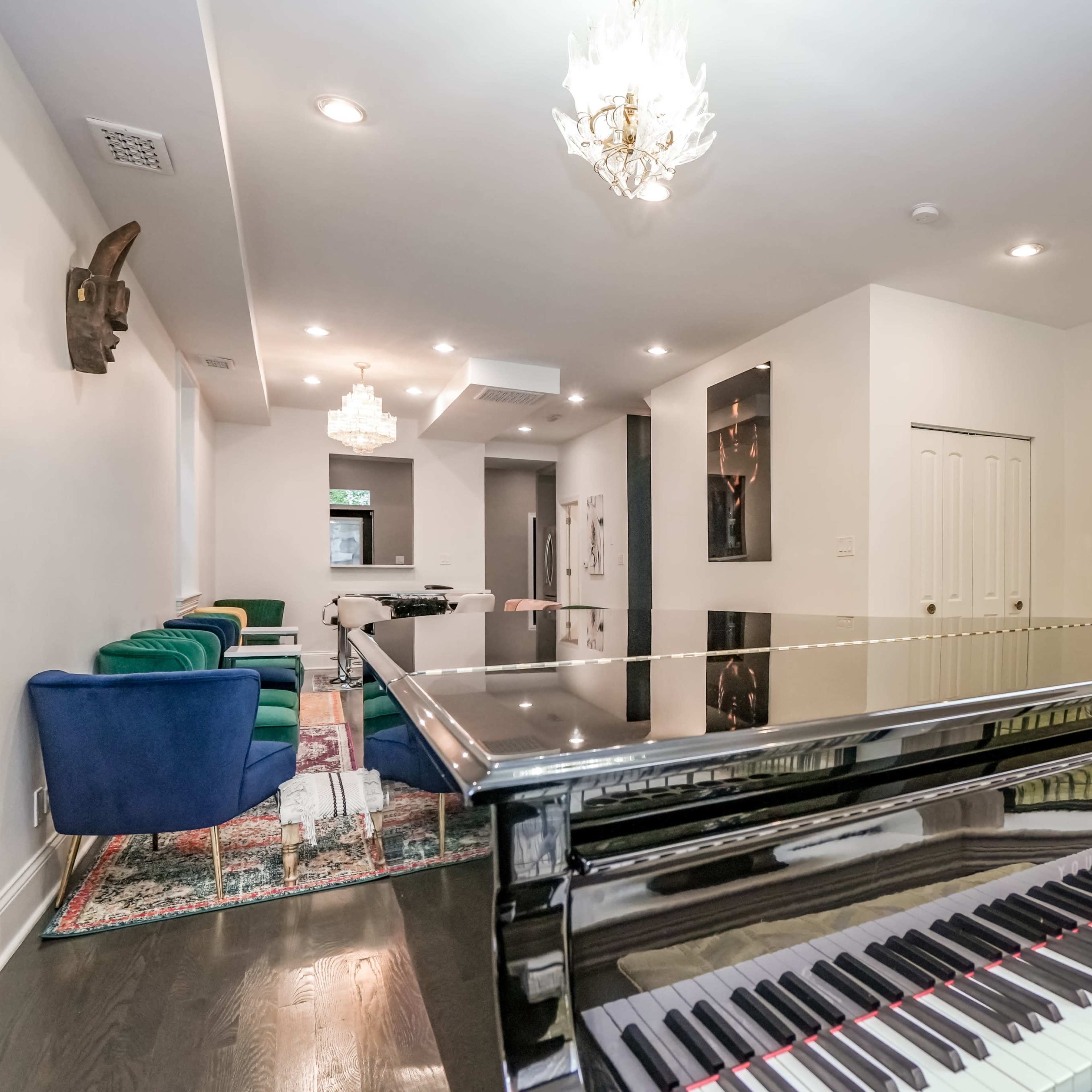 A black grand piano is showcased in the foreground, while a modern living space with green chairs and recessed lighting is visible in the background.