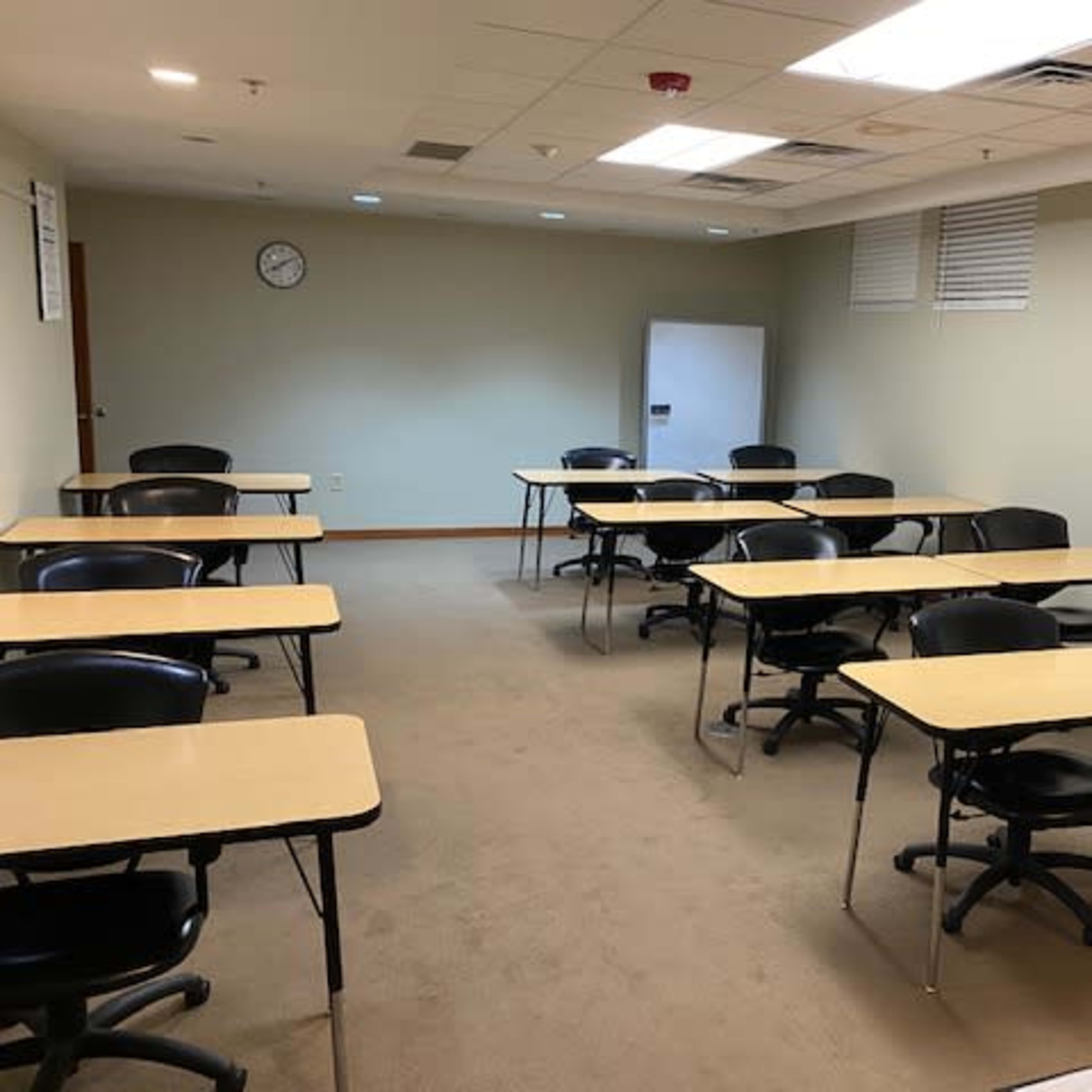 A classroom with several rows of tables and chairs arranged neatly on a carpeted floor, along with a wall clock and a refrigerator in the background.