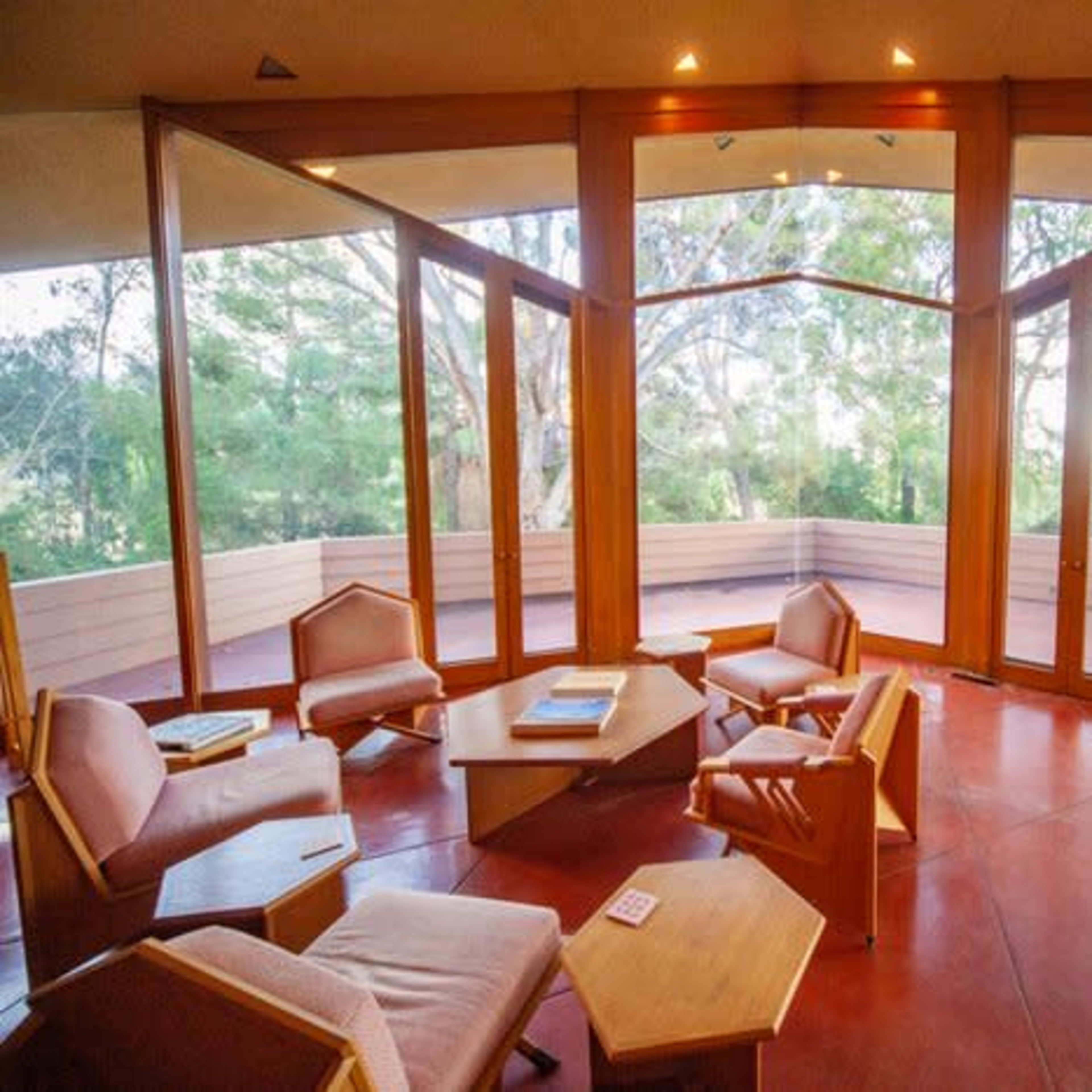 A modern interior space with large glass windows and wooden furniture, opening to an outdoor area surrounded by trees.
