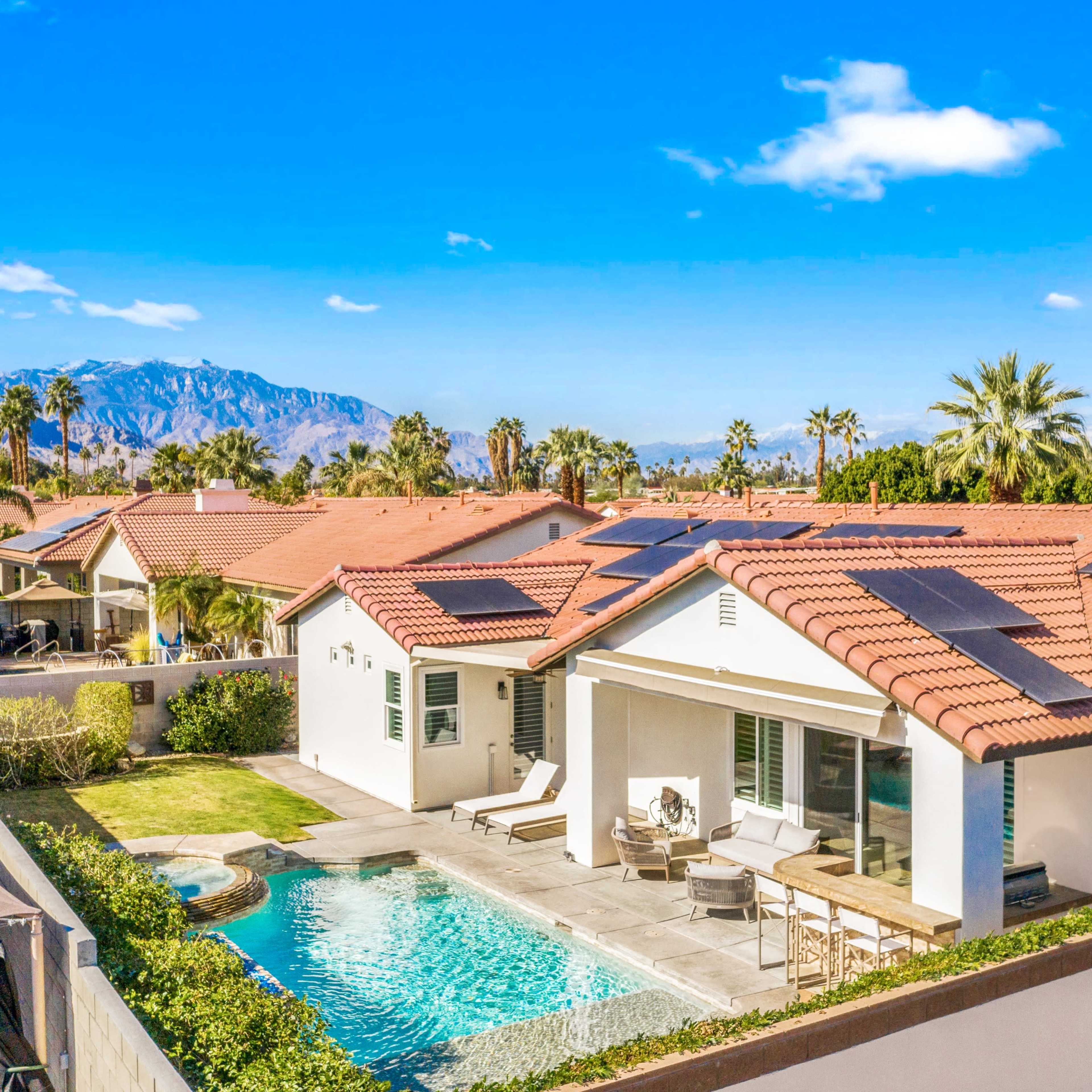 A modern house with a swimming pool and solar panels is surrounded by palm trees, with mountains visible in the background.