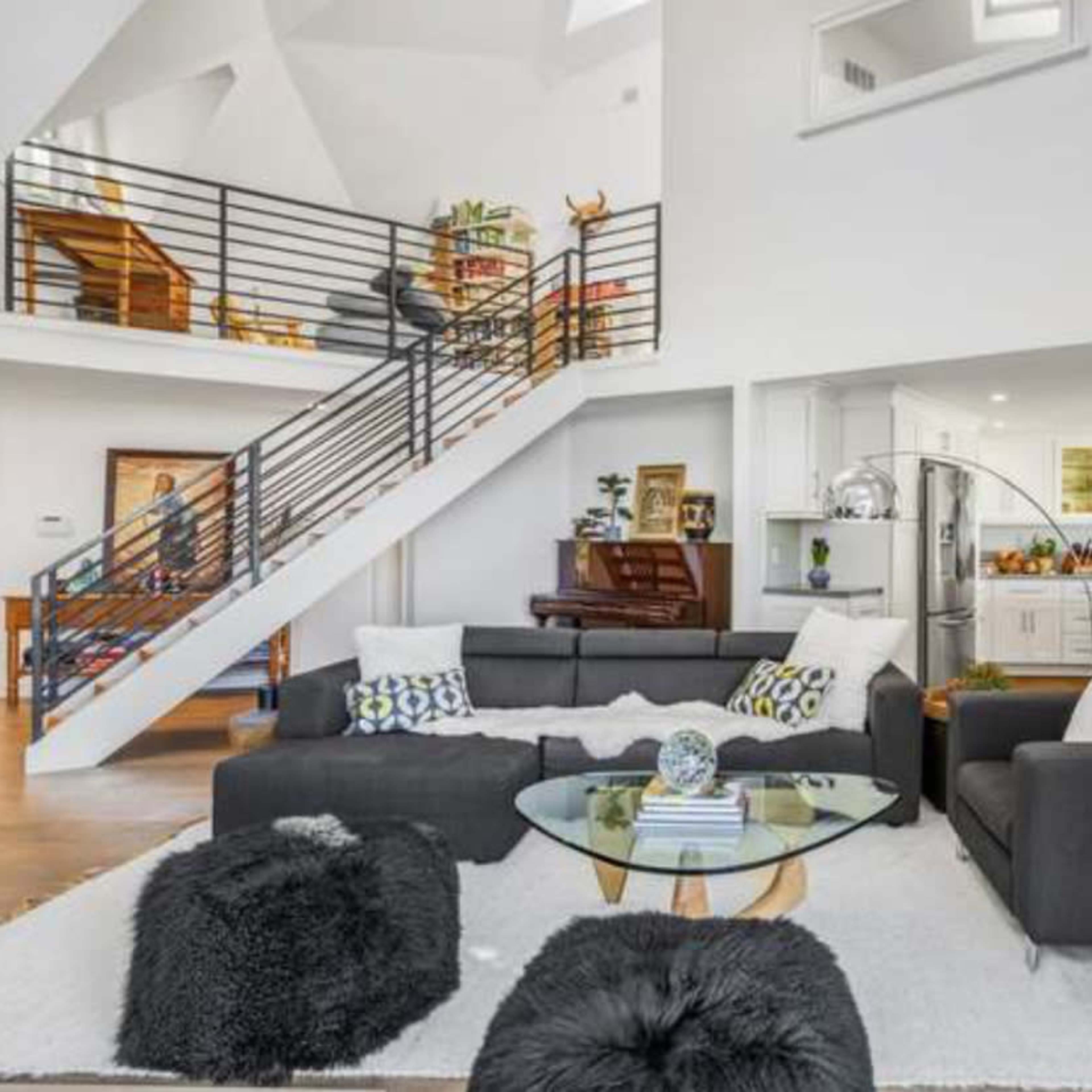 A modern living room with a gray sofa, a glass coffee table, and a staircase leading to an upper floor, adjacent to an open kitchen with a piano and dining area.