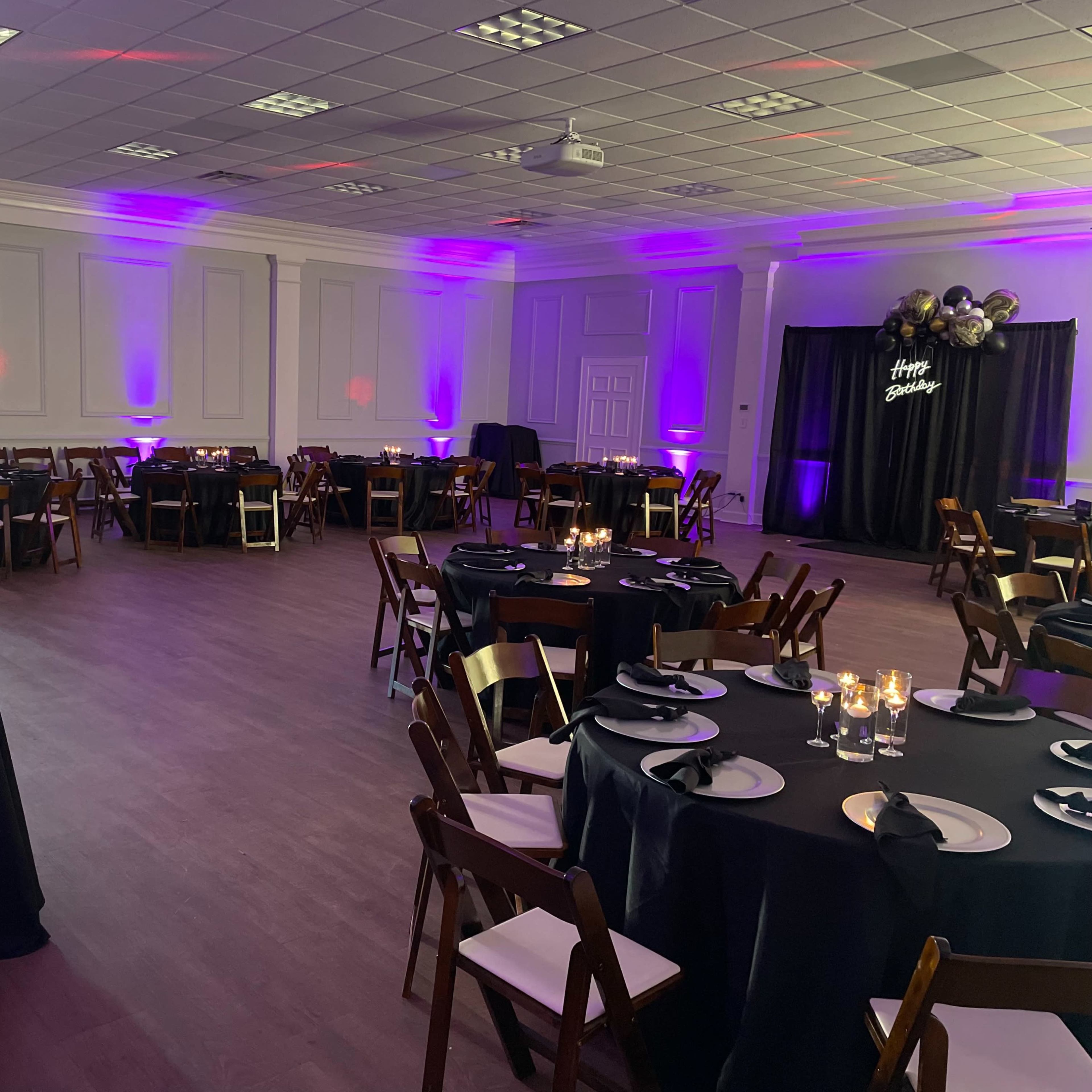 The room is set up for an event, featuring round tables with black tablecloths, wooden chairs, and purple lighting surrounding the space.