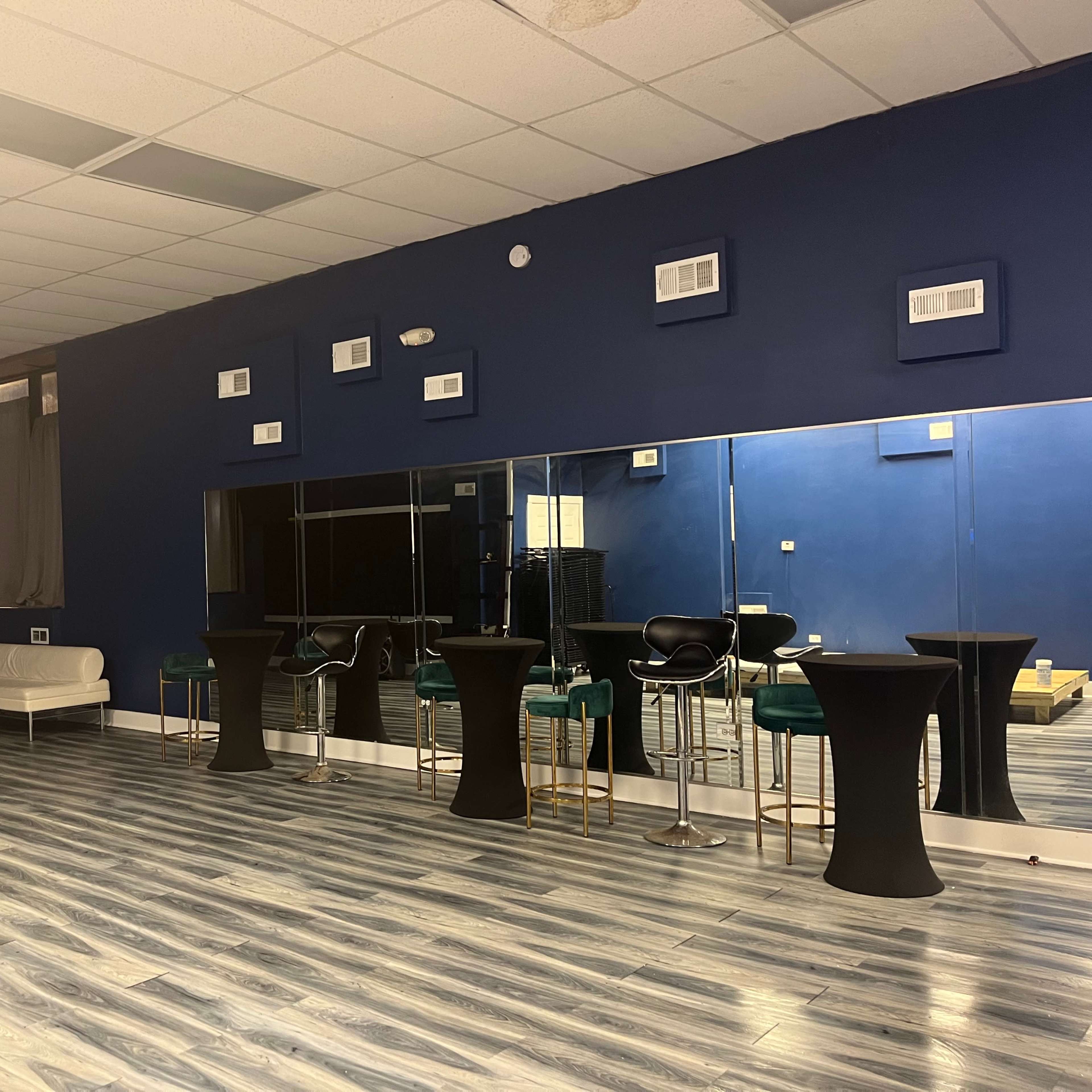 A spacious room with blue walls, wooden flooring, several tall black tables, and a large mirror reflecting the seating area.
