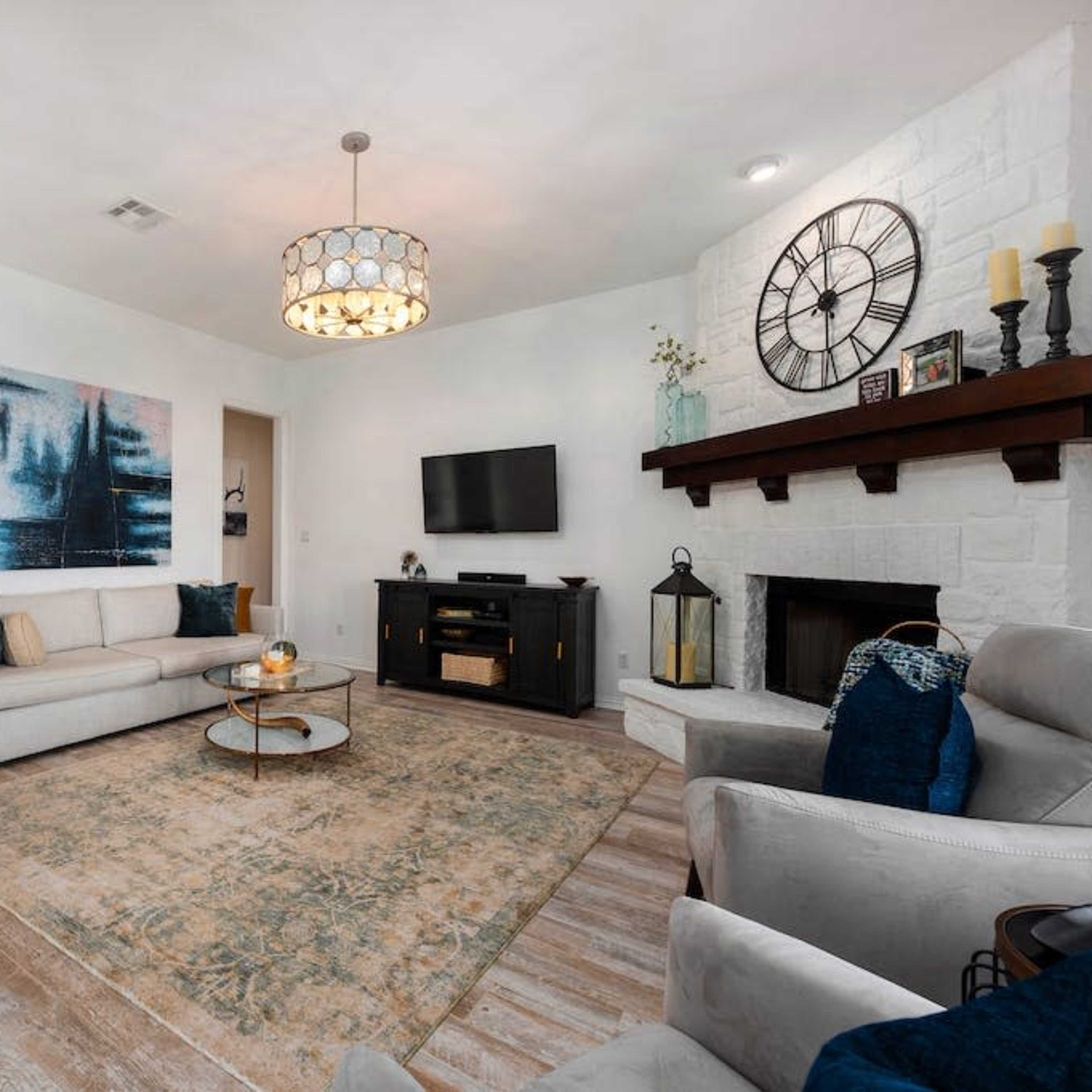 The living room features a white sofa, a round coffee table, a dark media console, and a stone fireplace, with a large wall art piece and a decorative chandelier overhead.