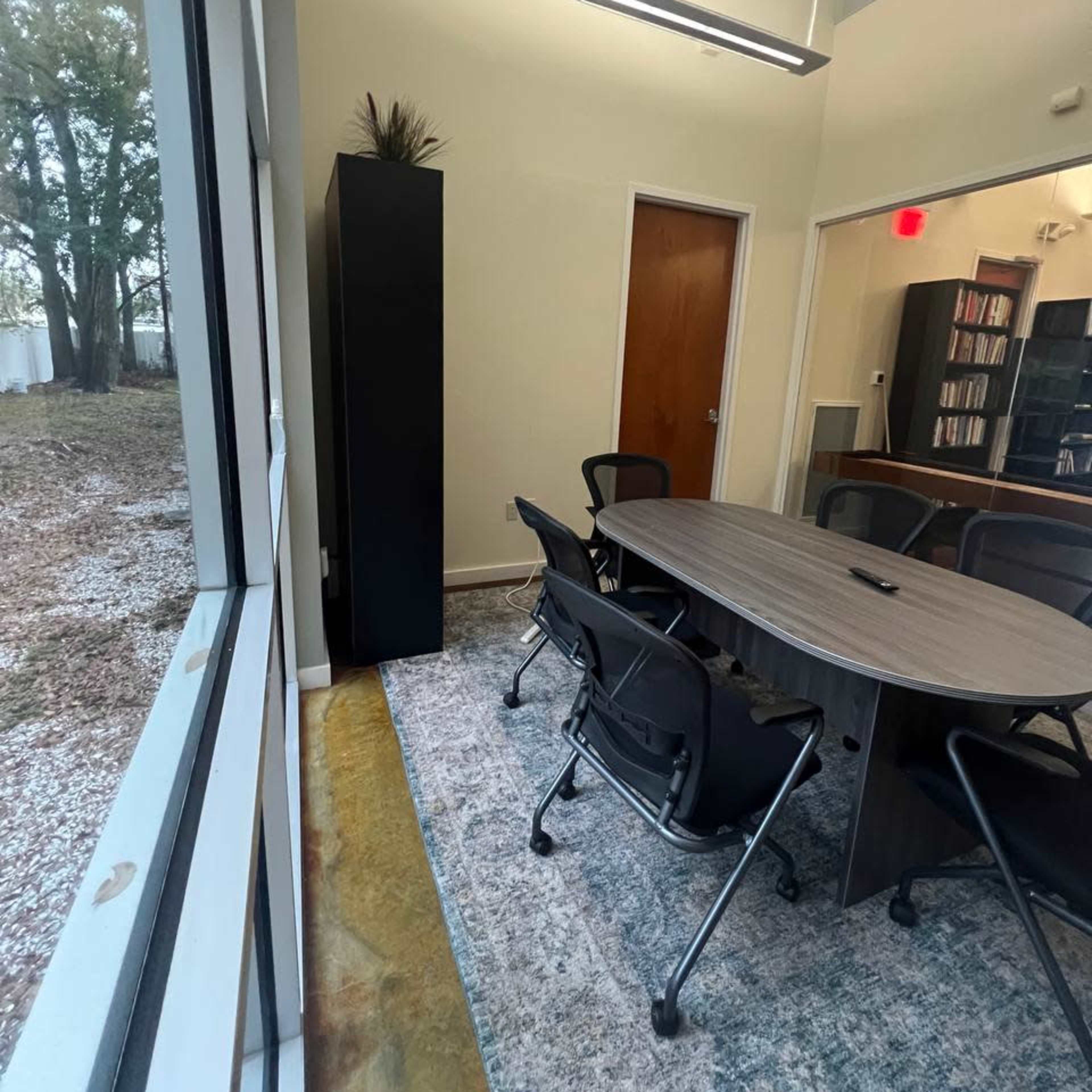 A conference room features a round table surrounded by chairs, with a view of trees through large windows.
