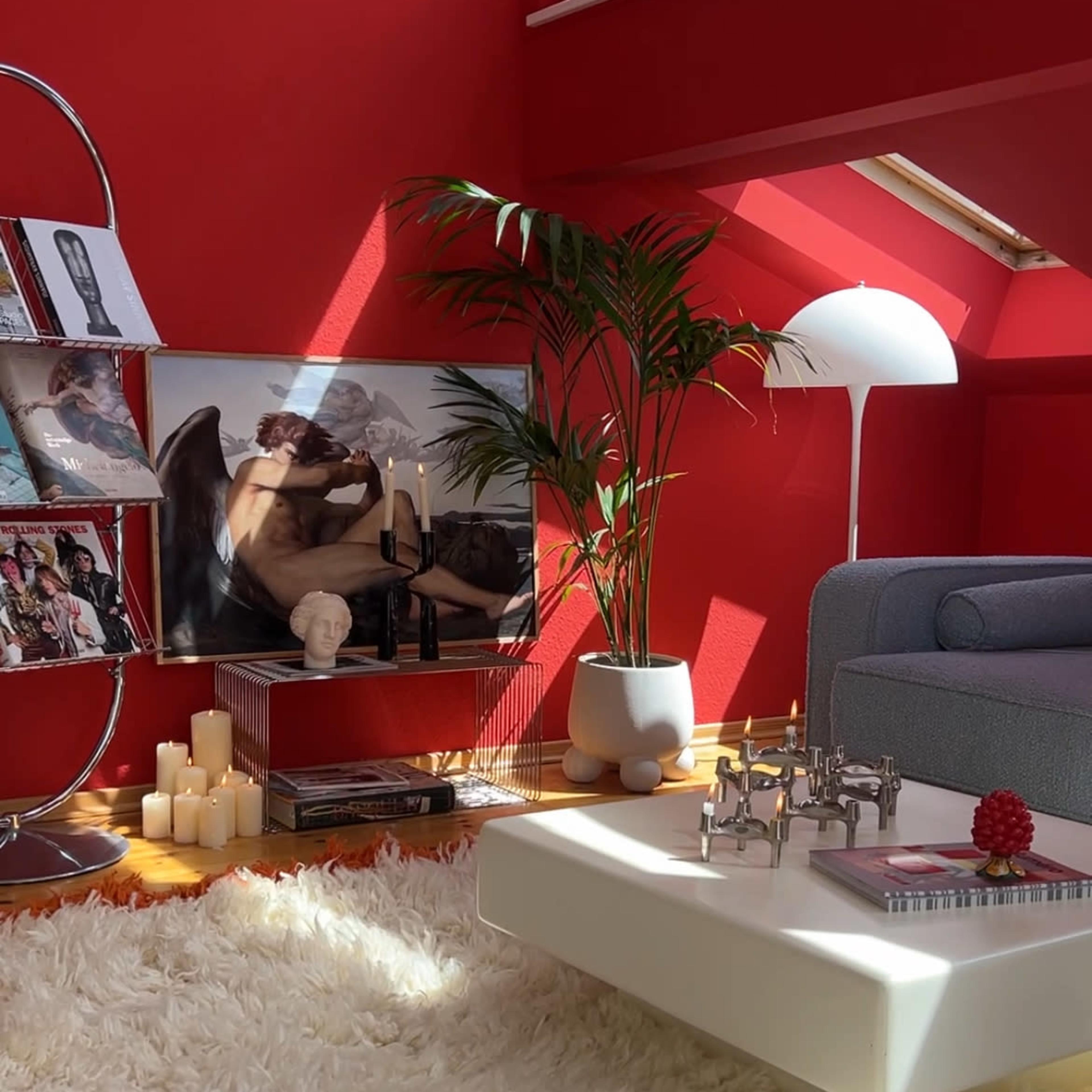 The scene depicts a modern living room with a red wall, a couch, a coffee table, and candles arranged on a fluffy rug.