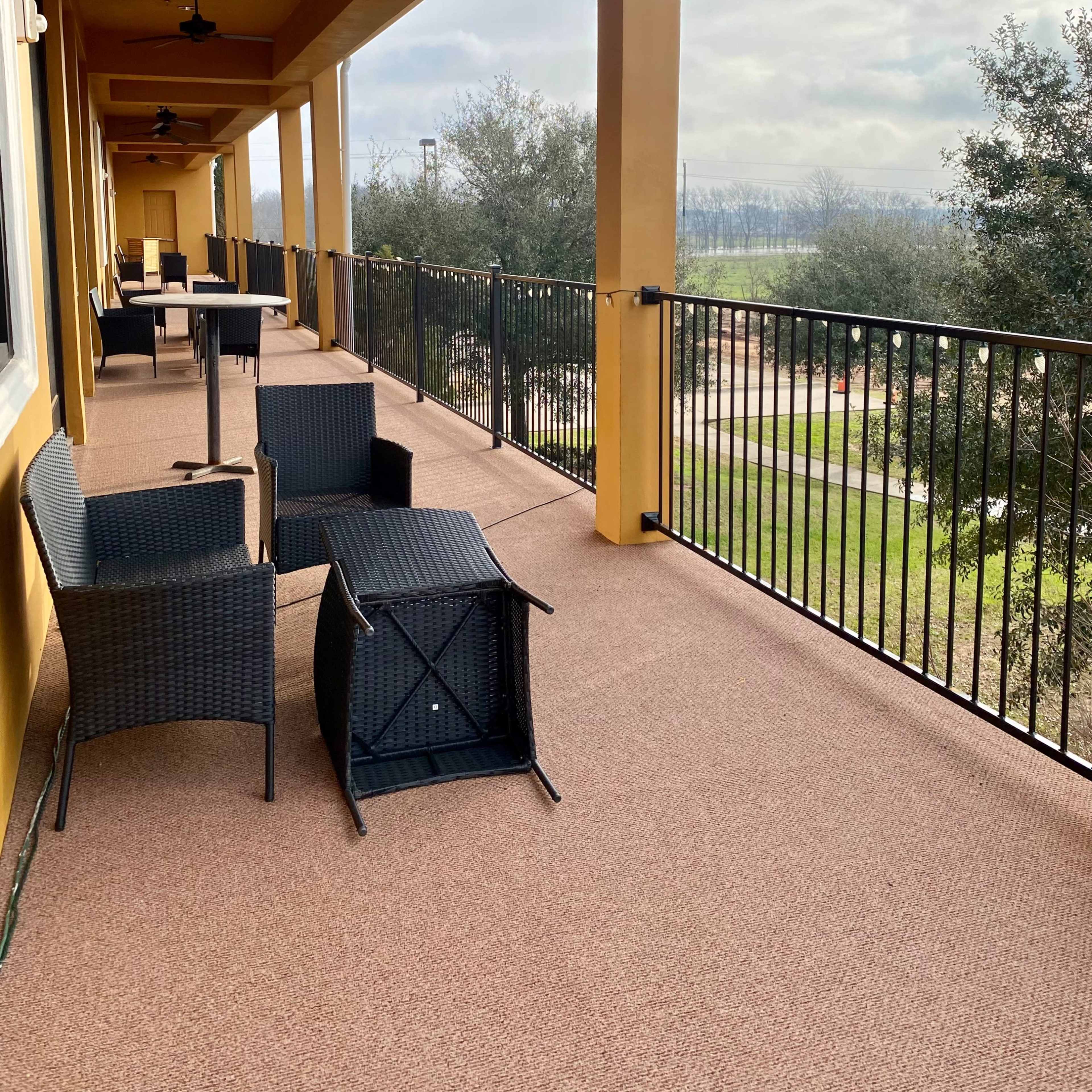 A covered balcony with black wicker furniture and a view of a grassy area and trees in the distance.