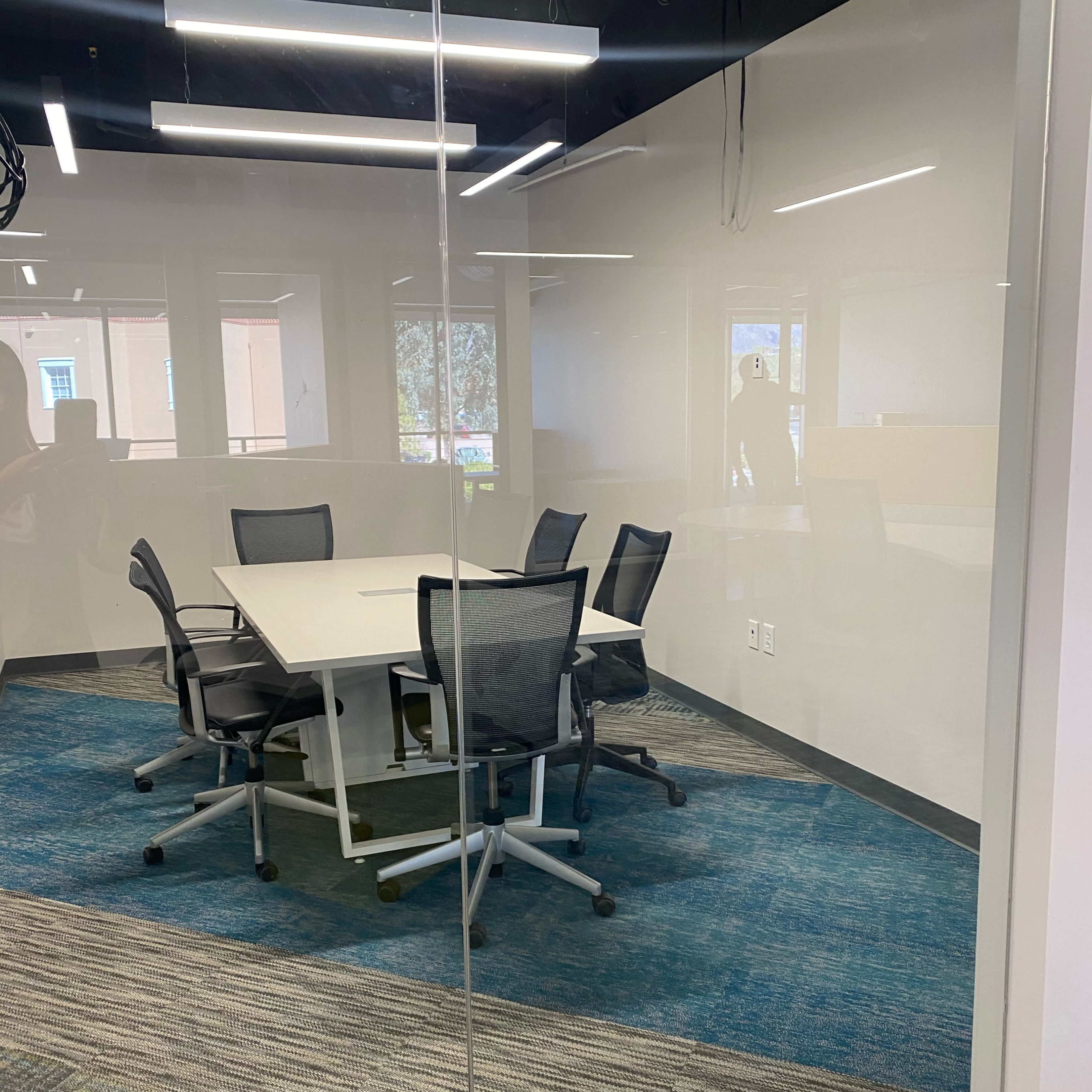A glass-walled conference room features a round table surrounded by six office chairs on a patterned carpet.