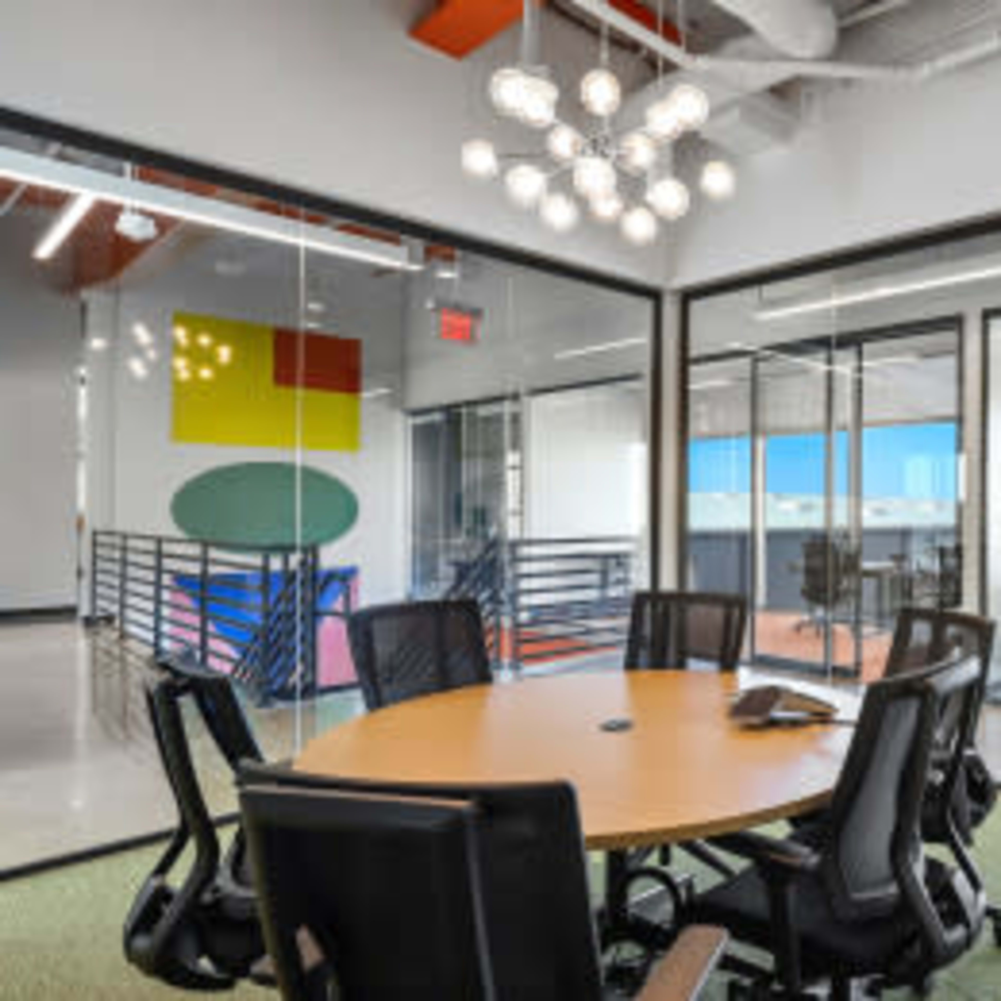 A modern conference room features a round table surrounded by black office chairs, with glass walls providing a view of adjacent work areas.