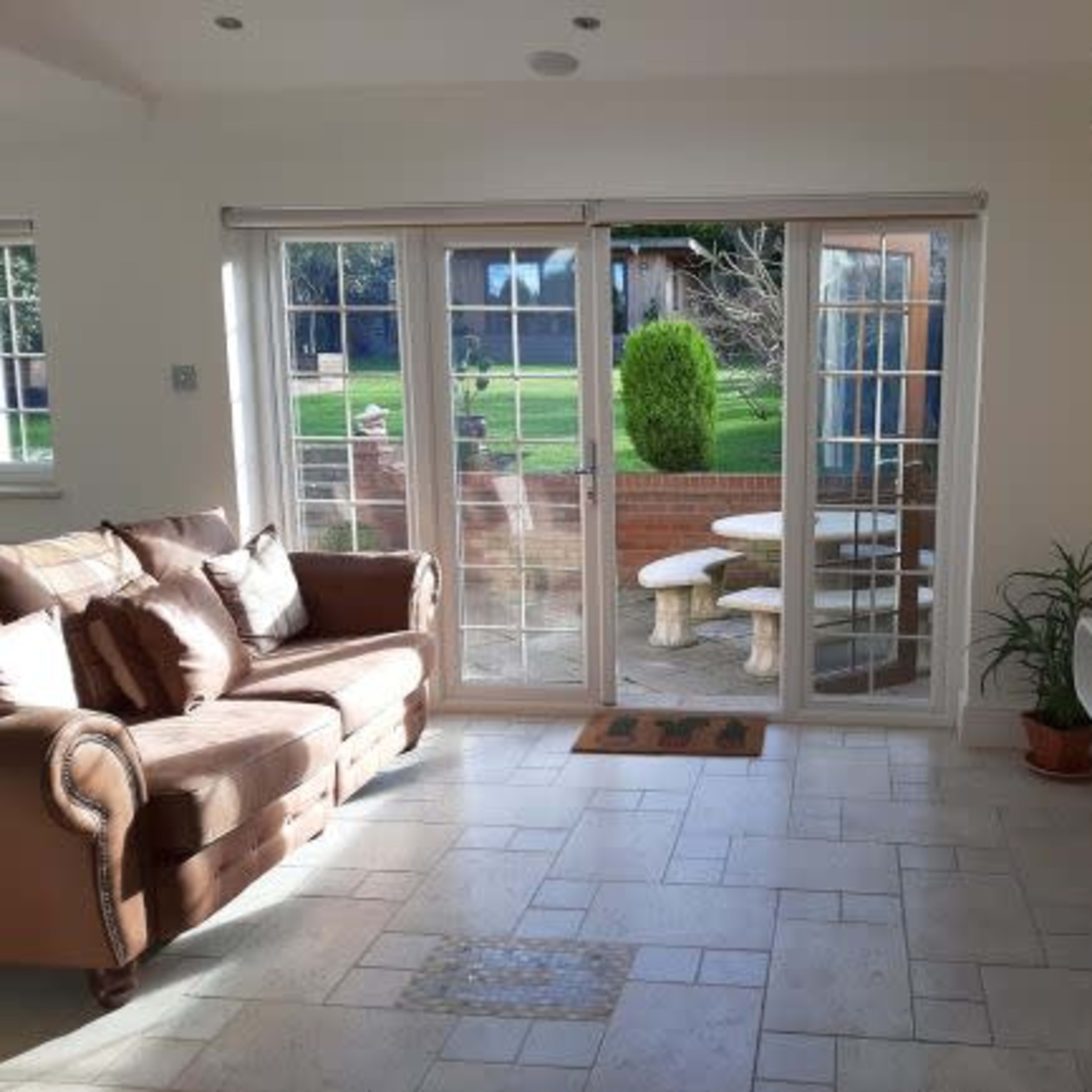 The living room features a brown sofa, a wall-mounted TV, and large glass doors leading to a garden area with a stone table and benches.