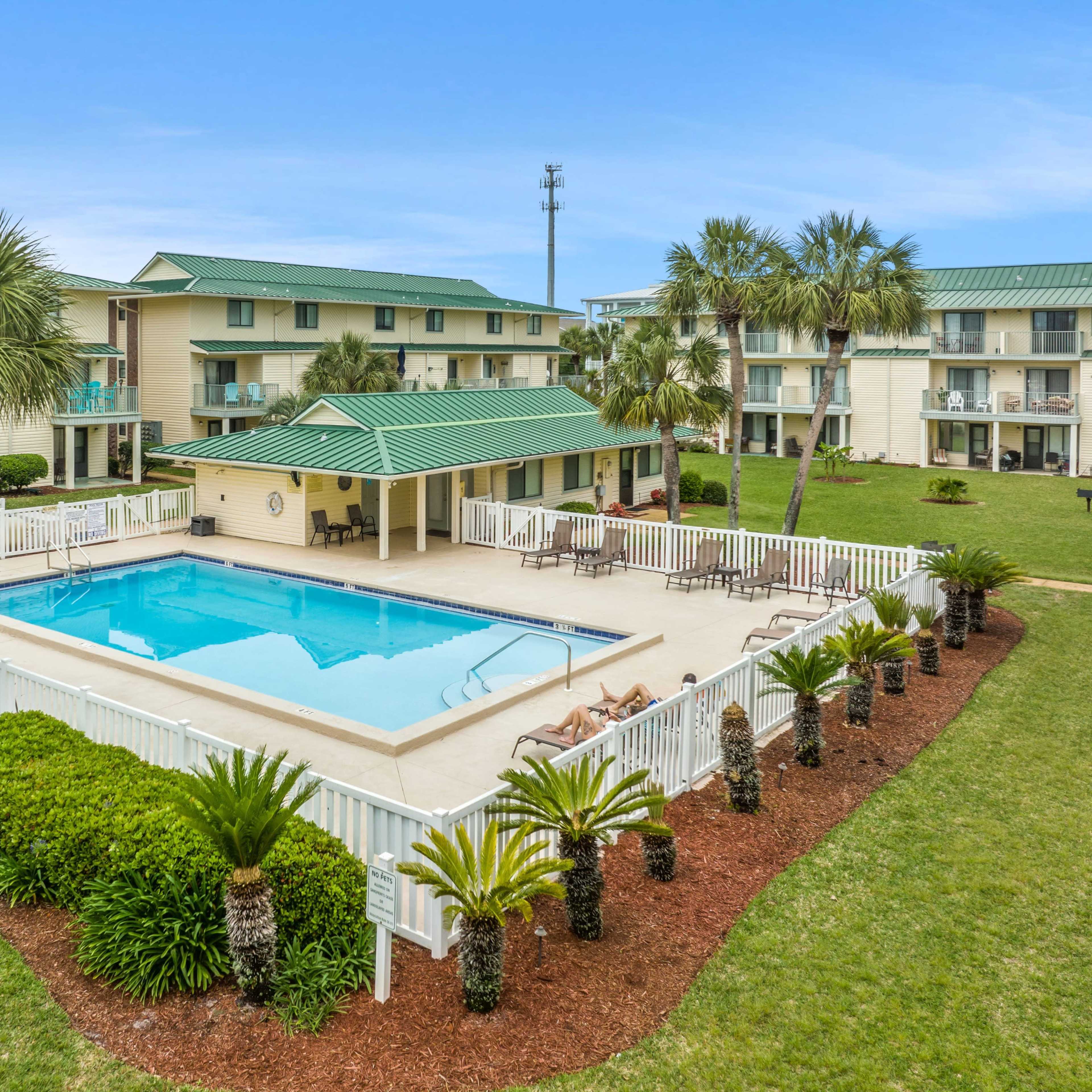 A well-maintained swimming pool surrounded by a white fence, palm trees, and green landscaping at a resort-like property.