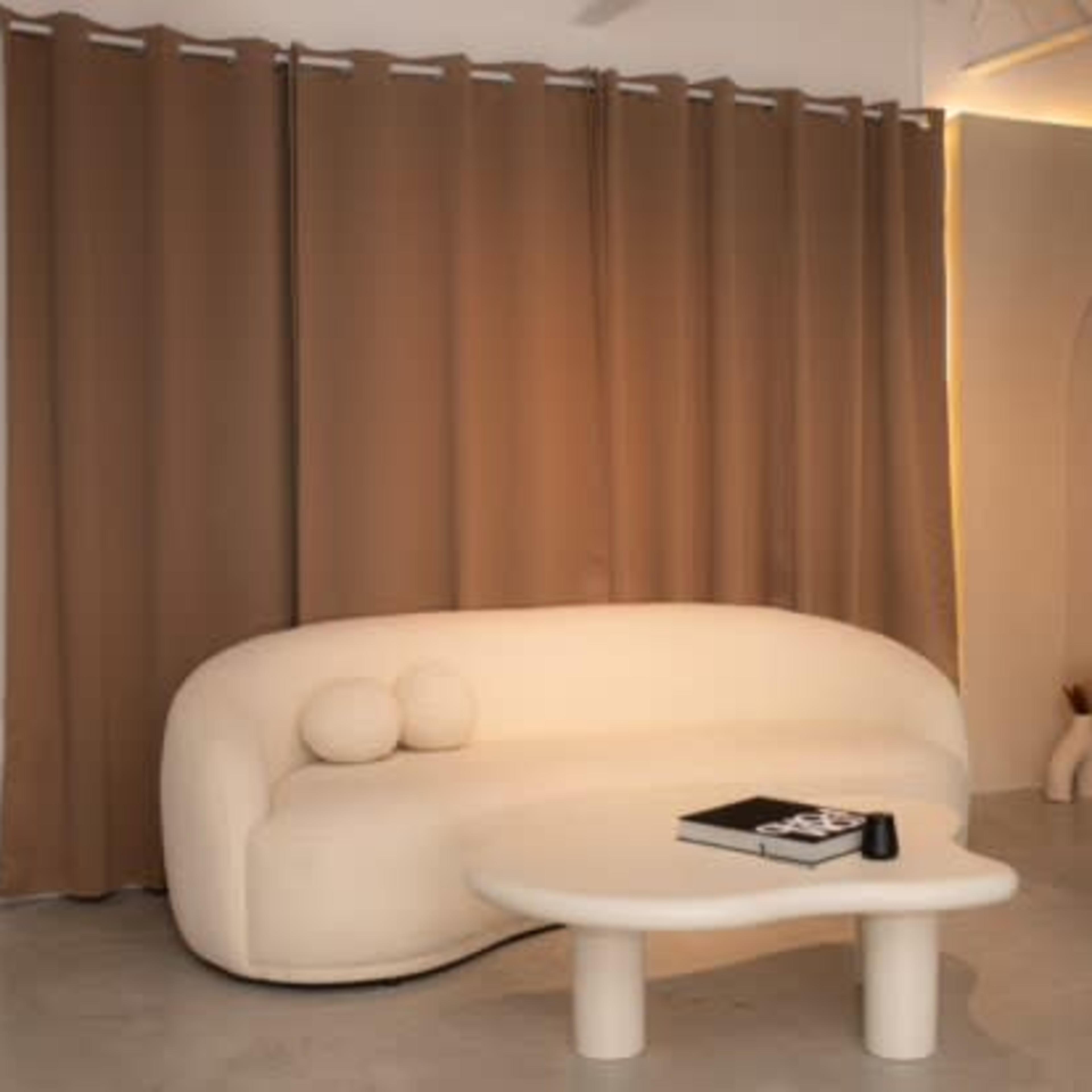 A minimalistic living space featuring a curved white sofa, a round coffee table, and brown curtains, with soft lighting and decorative elements in the background.