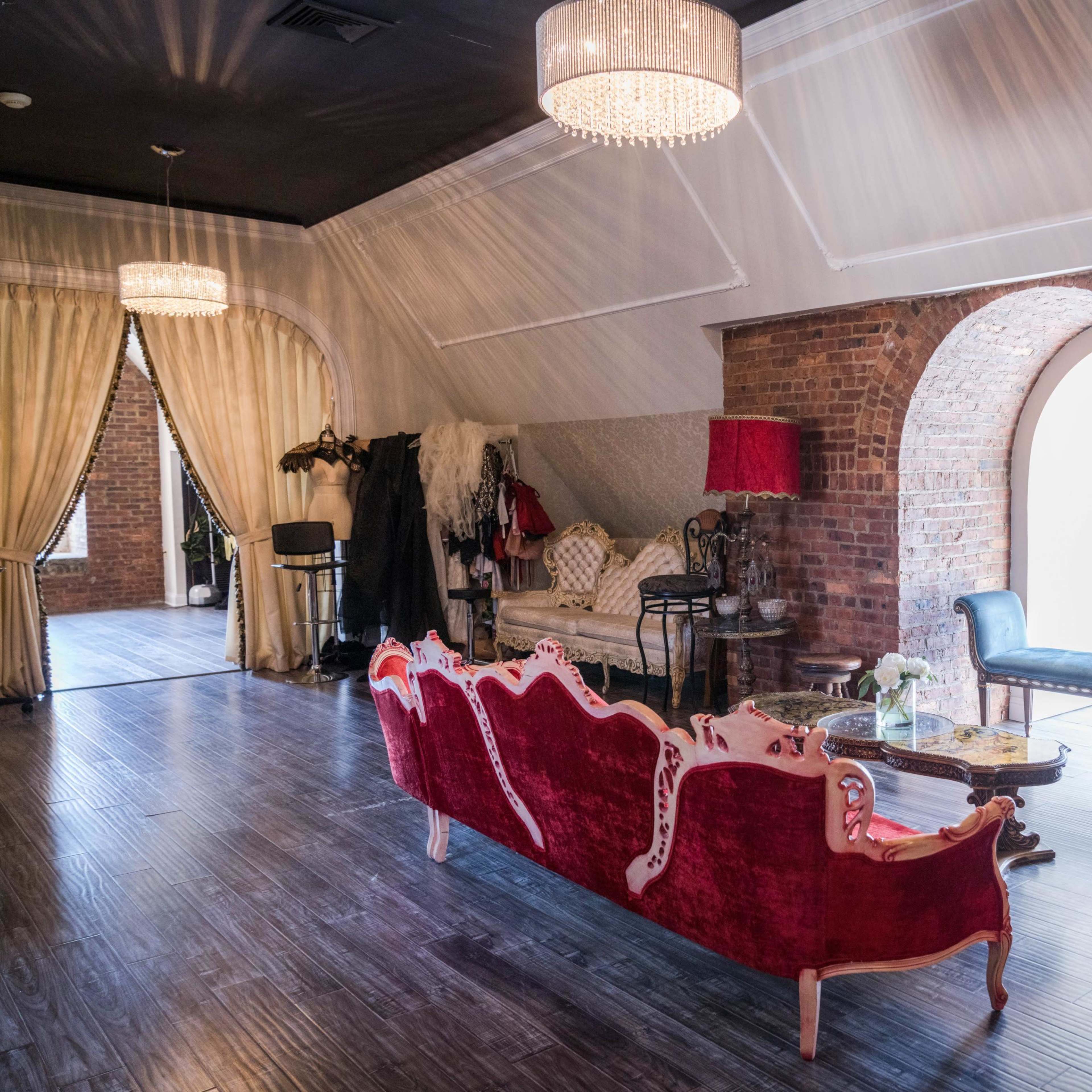 The room features a red velvet sofa and armchair arranged near a brick wall with arched windows, complemented by elegant lighting fixtures and decorative elements.