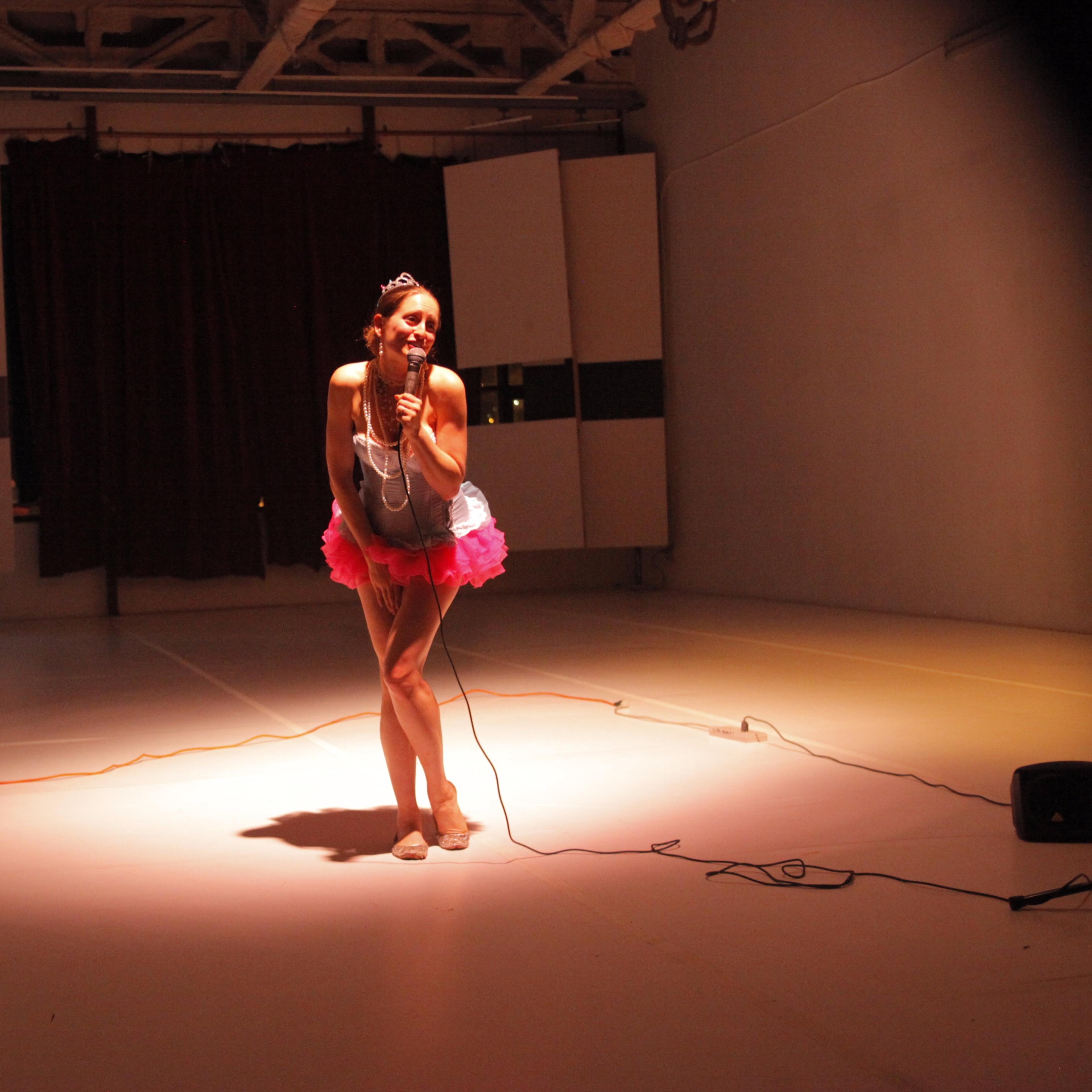 A performer in a colorful costume stands on stage with a microphone in a dimly lit room.