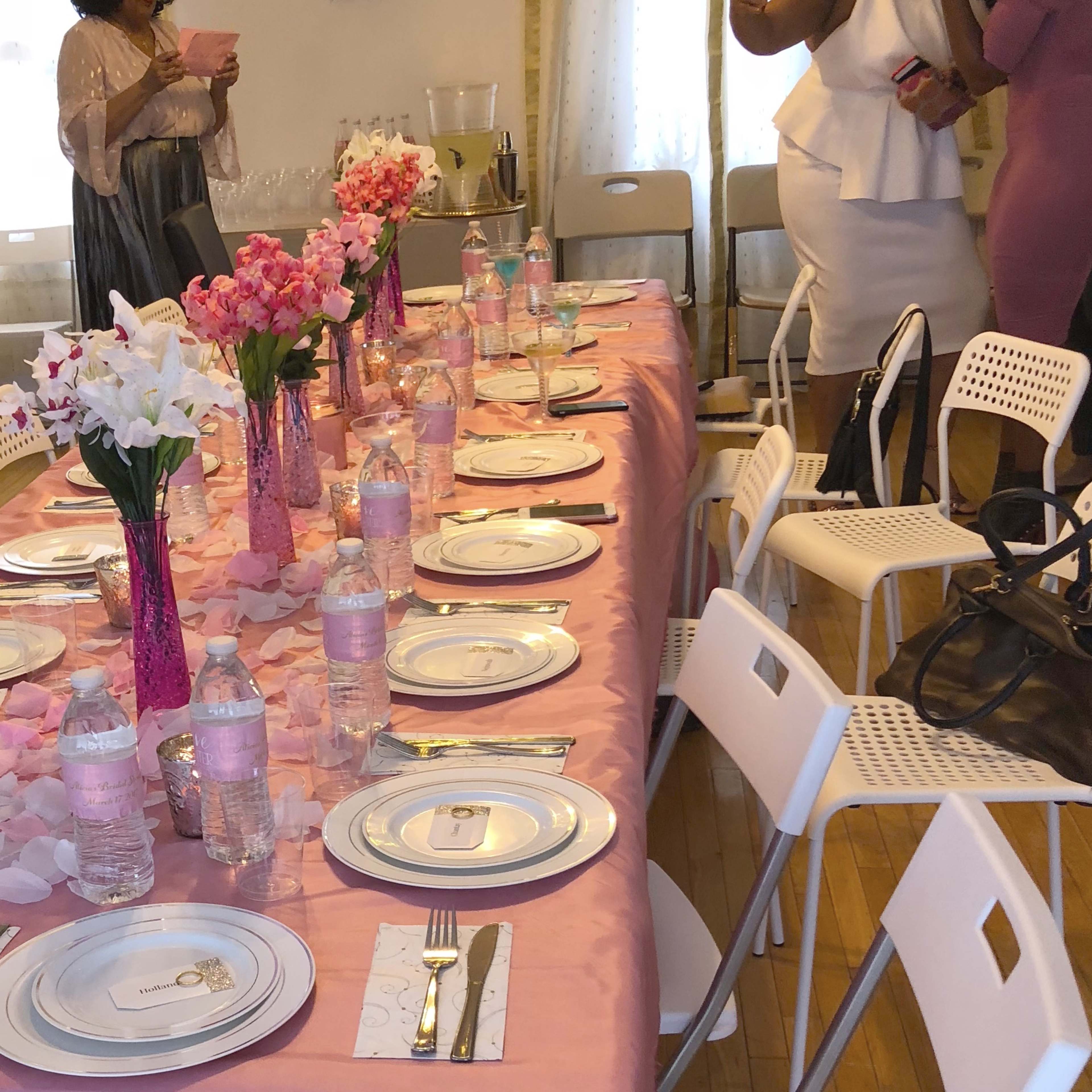 A long table is set with pink tablecloths, floral centerpieces, and water bottles, surrounded by guests at a gathering.