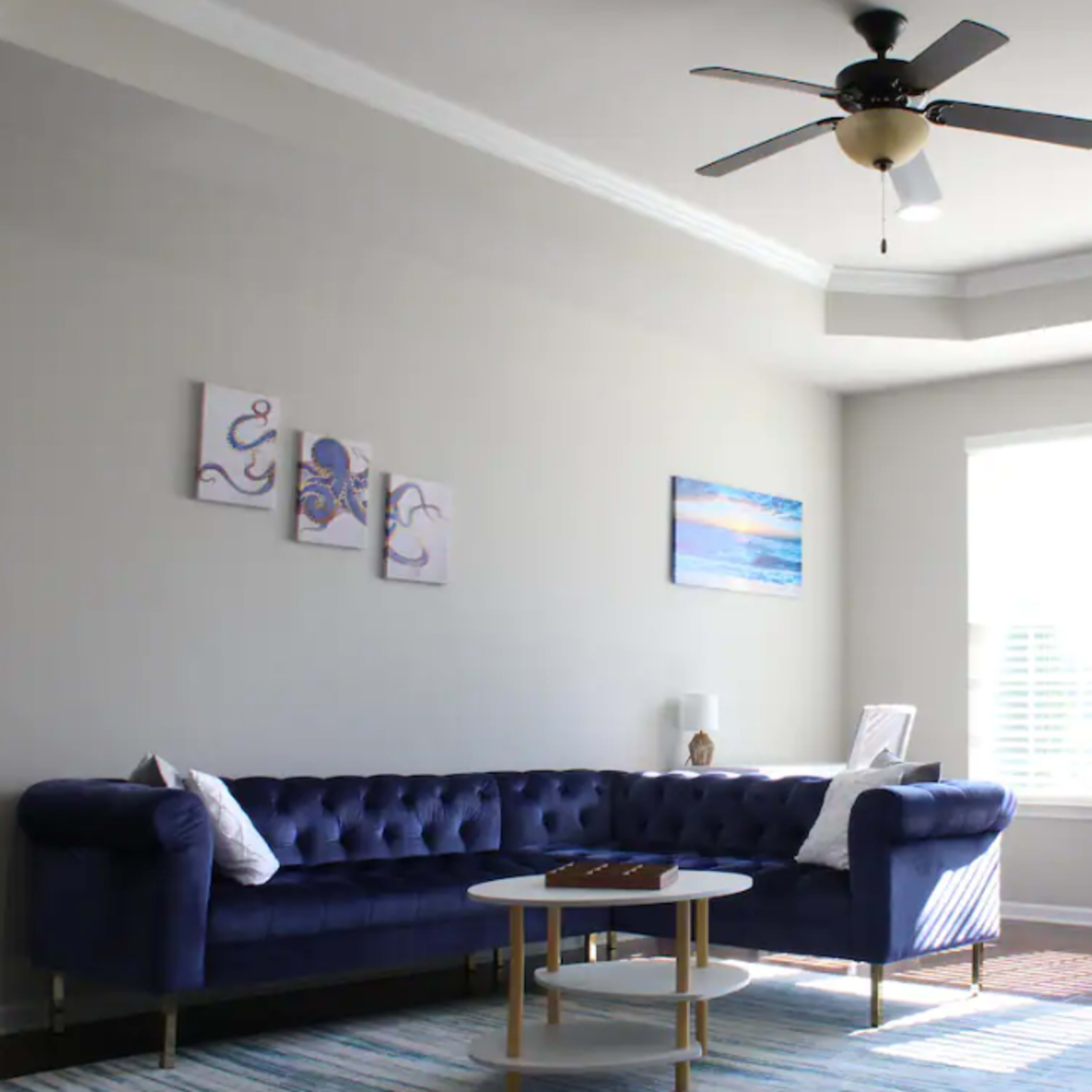 A blue tufted sofa is positioned in a well-lit living room with large windows and a ceiling fan, accompanied by a coffee table and wall art.