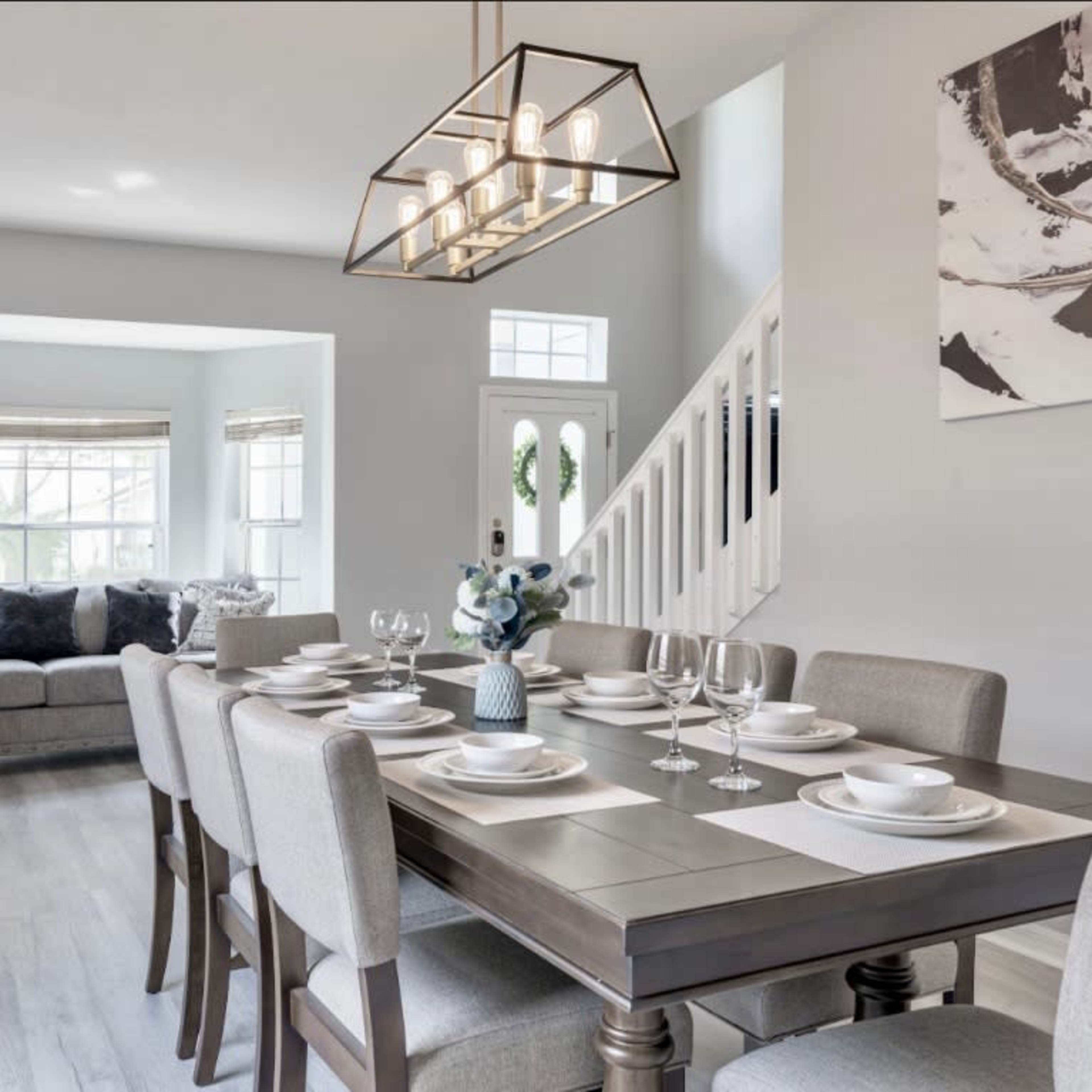 A modern dining room features a large table set for dinner, surrounded by beige chairs, with a cozy seating area visible in the background.