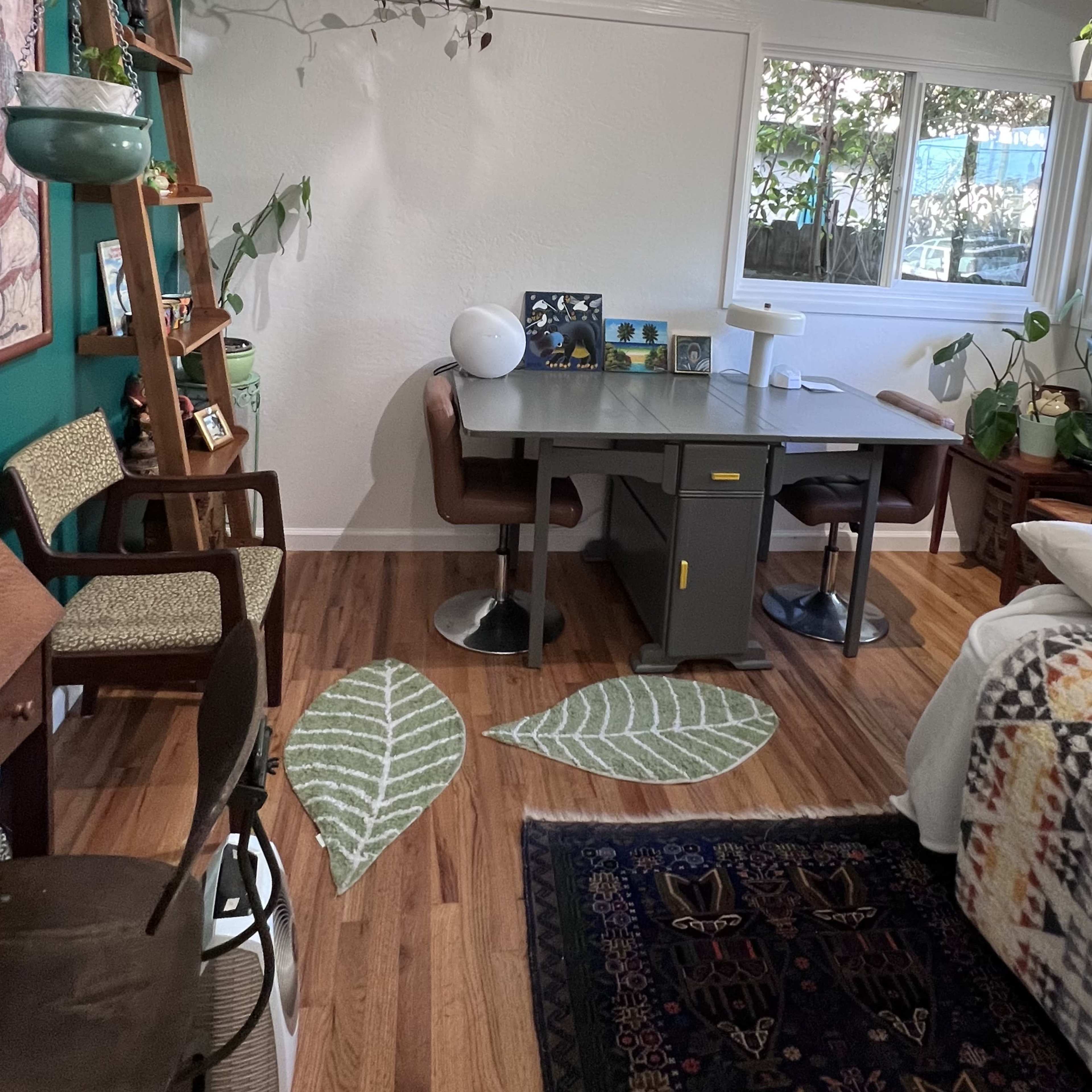A cozy indoor workspace featuring a desk with two chairs, surrounded by plants and a window that lets in natural light.